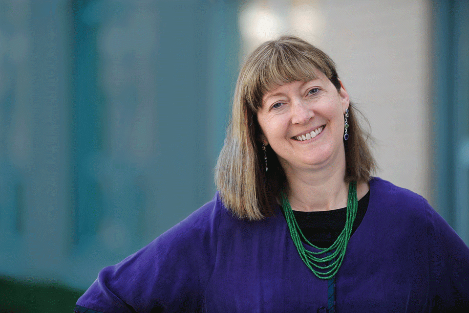A smiling middle-aged woman with shoulder-length brown hair, wearing a purple top, a green beaded necklace, and earrings, standing outdoors in front of a blurred background.