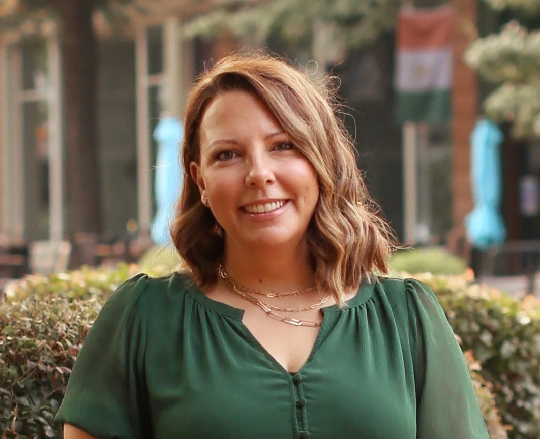 A woman with shoulder-length, wavy brown hair smiling outdoors, wearing a green blouse and layered necklaces, with a background of bushes, buildings, and a flag.