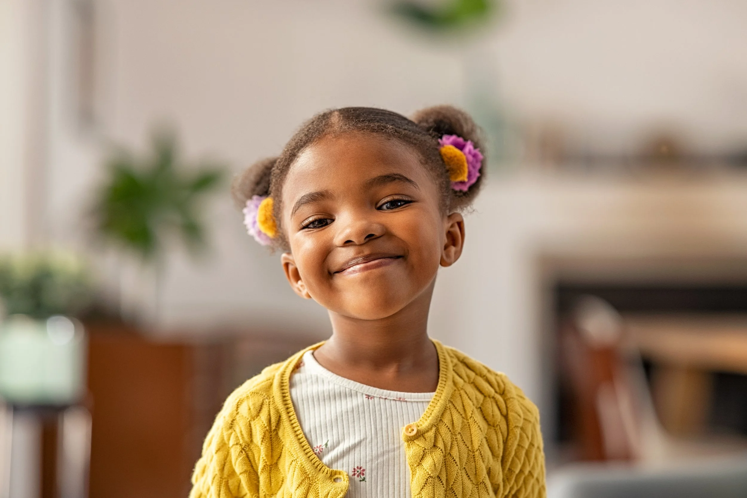 A young girl with a smile and colorful flower hair accessories, wearing a yellow cardigan, standing indoors with a blurred background.
