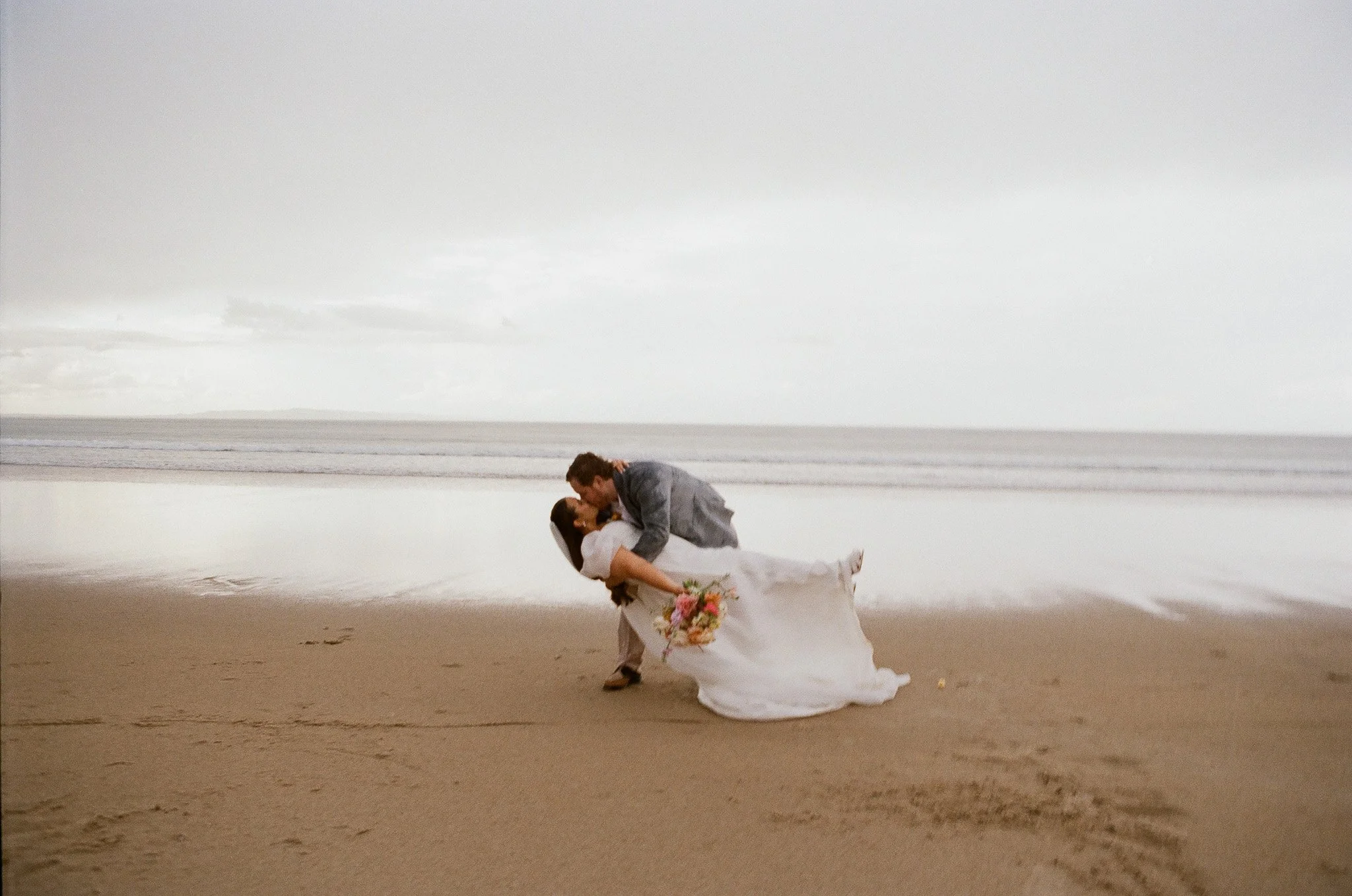 A newlywed couple on the beach, with the groom dipping the bride as they kiss, the bride holding a bouquet of flowers, with the ocean and cloudy sky in the background.