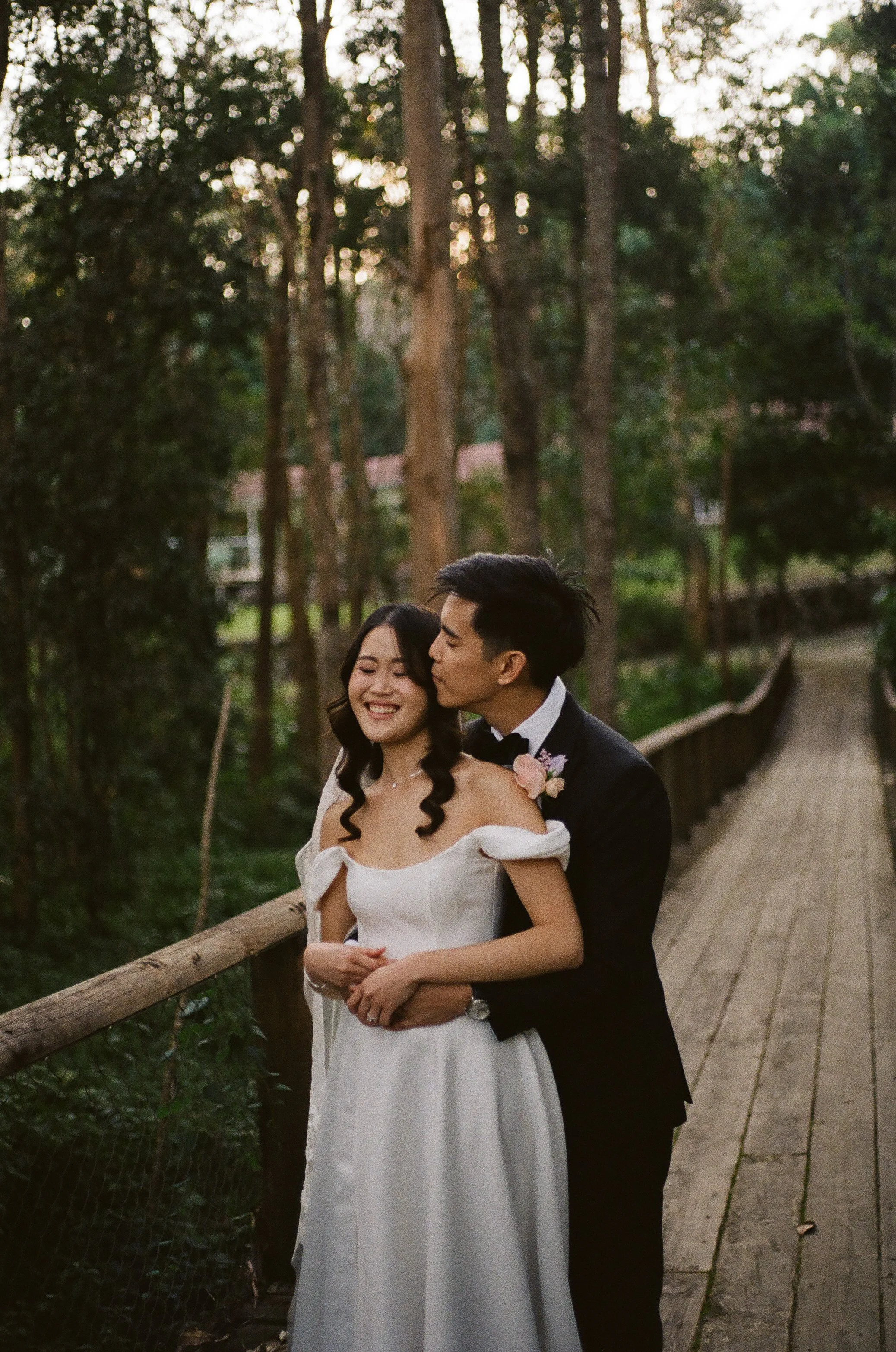 A happy bride and groom on their wedding day, standing on a wooden pathway in a forest, with the groom kissing the bride's cheek and the bride smiling with eyes closed.