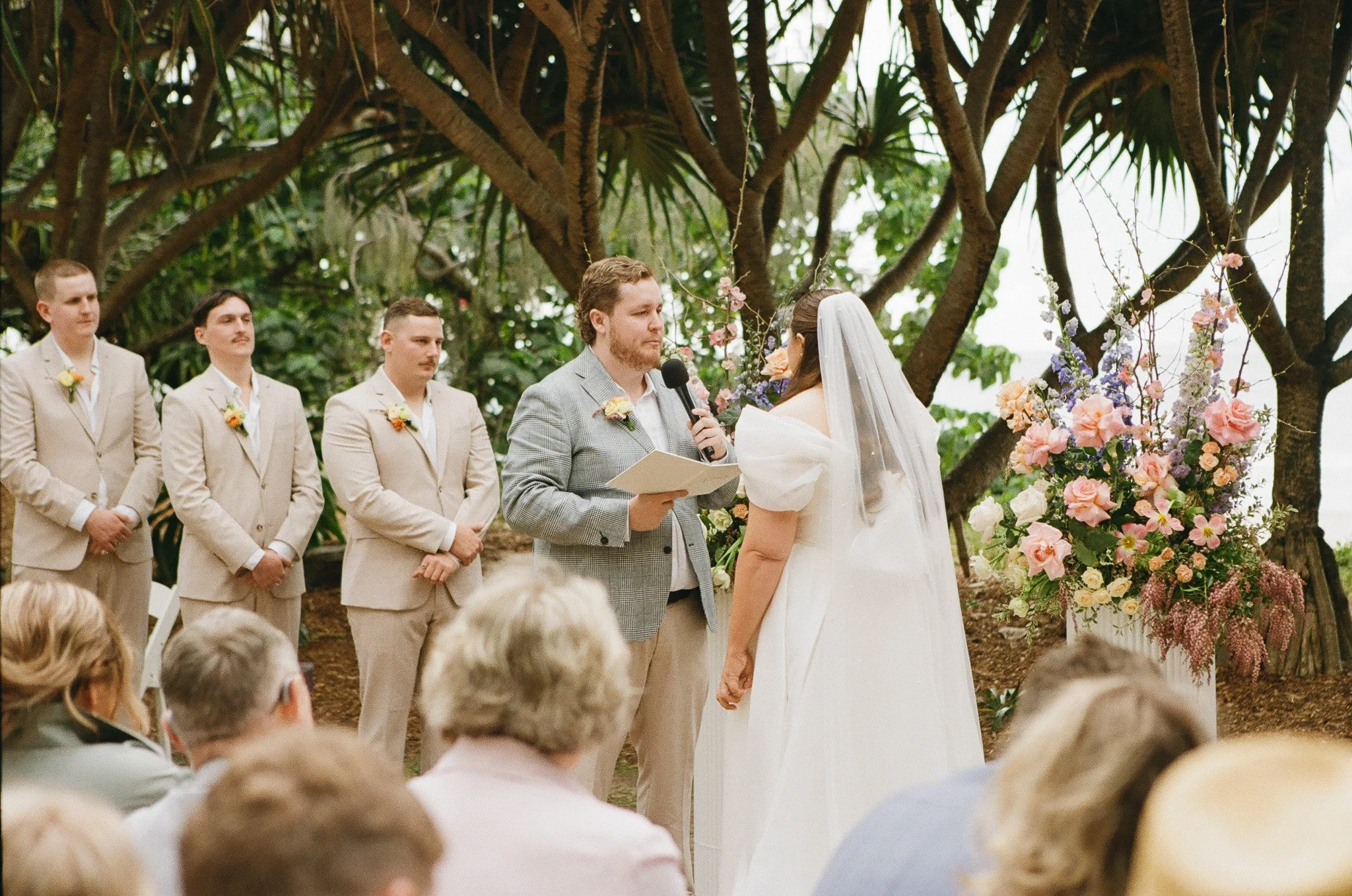 A wedding ceremony outdoors with a couple exchanging vows, a man in a gray suit holding a microphone and paper, and four groomsmen in beige suits standing to the side, amidst large trees and floral arrangements.