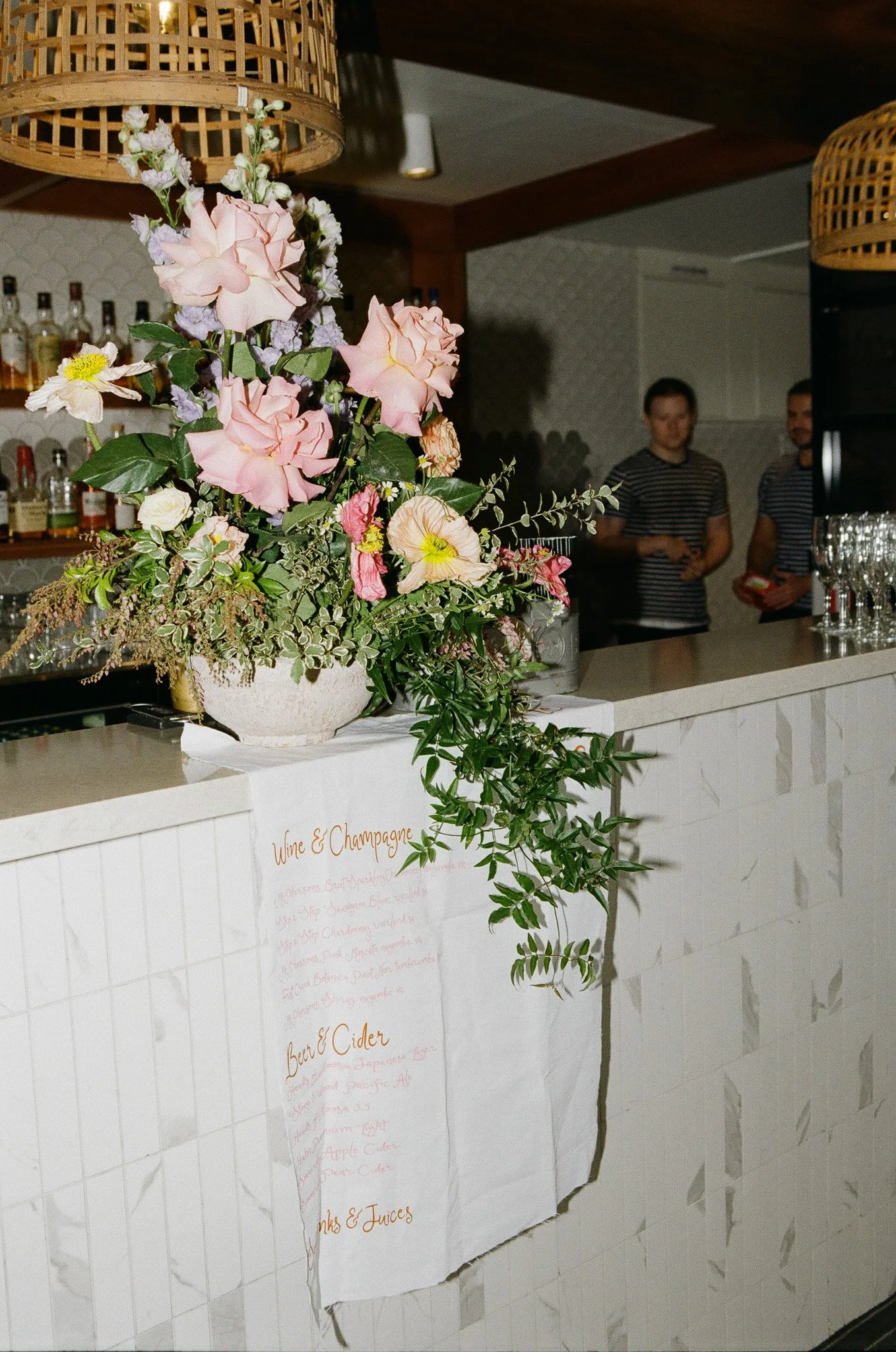 A floral arrangement with pink roses, white and purple smaller flowers, and green foliage on a white tiled bar with a menu sign. Two men in striped shirts stand in the background.