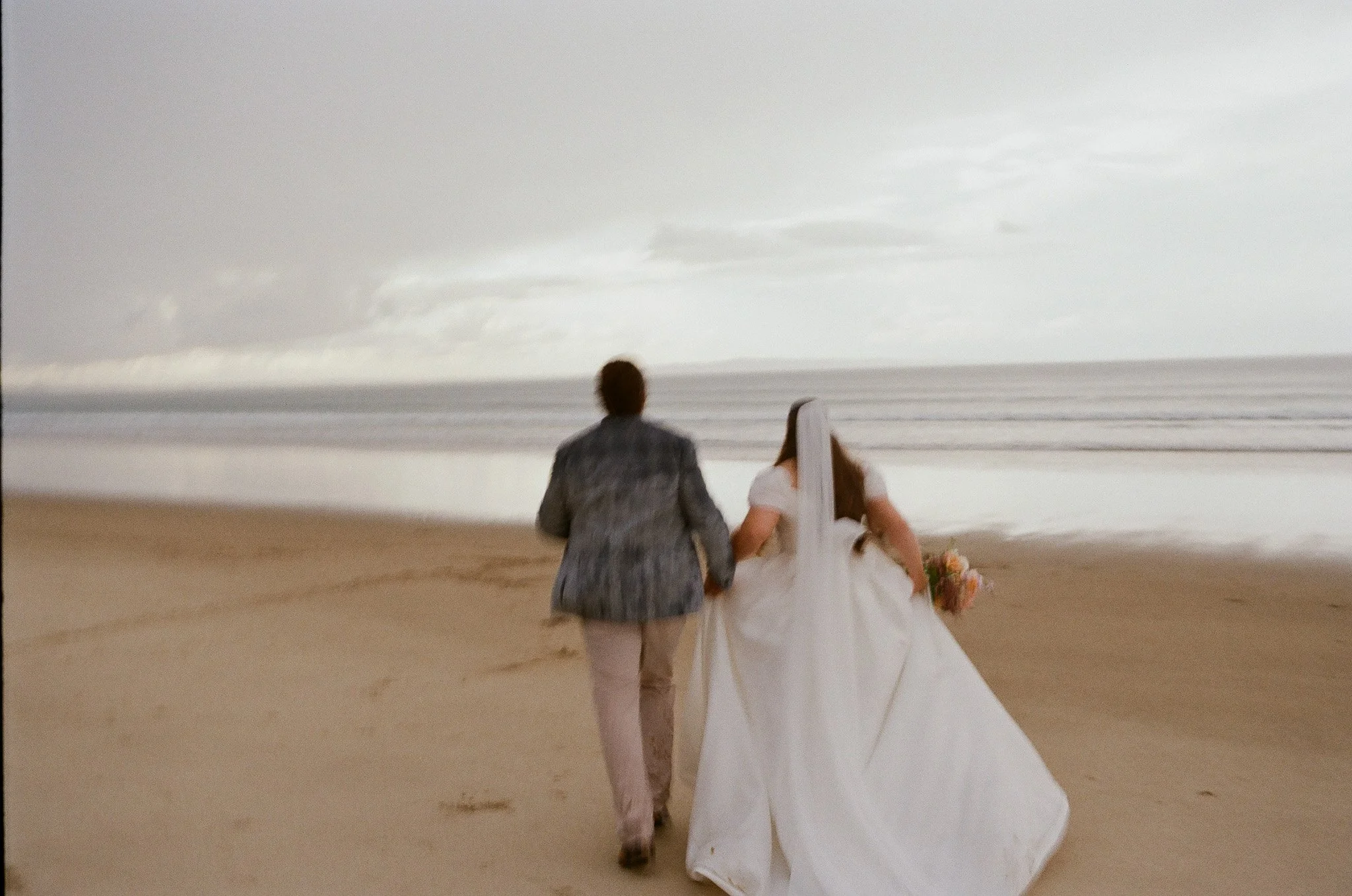 A bride and groom holding hands and walking on the beach, with the ocean and it cloudy sky in the background.