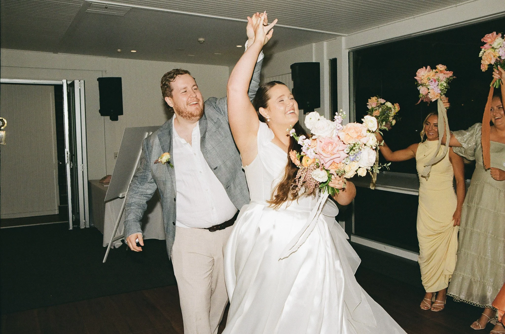 Happy bride holding a large bouquet of pink and white flowers, with a groom raising his hand in celebration, during a wedding reception with friends in the background.