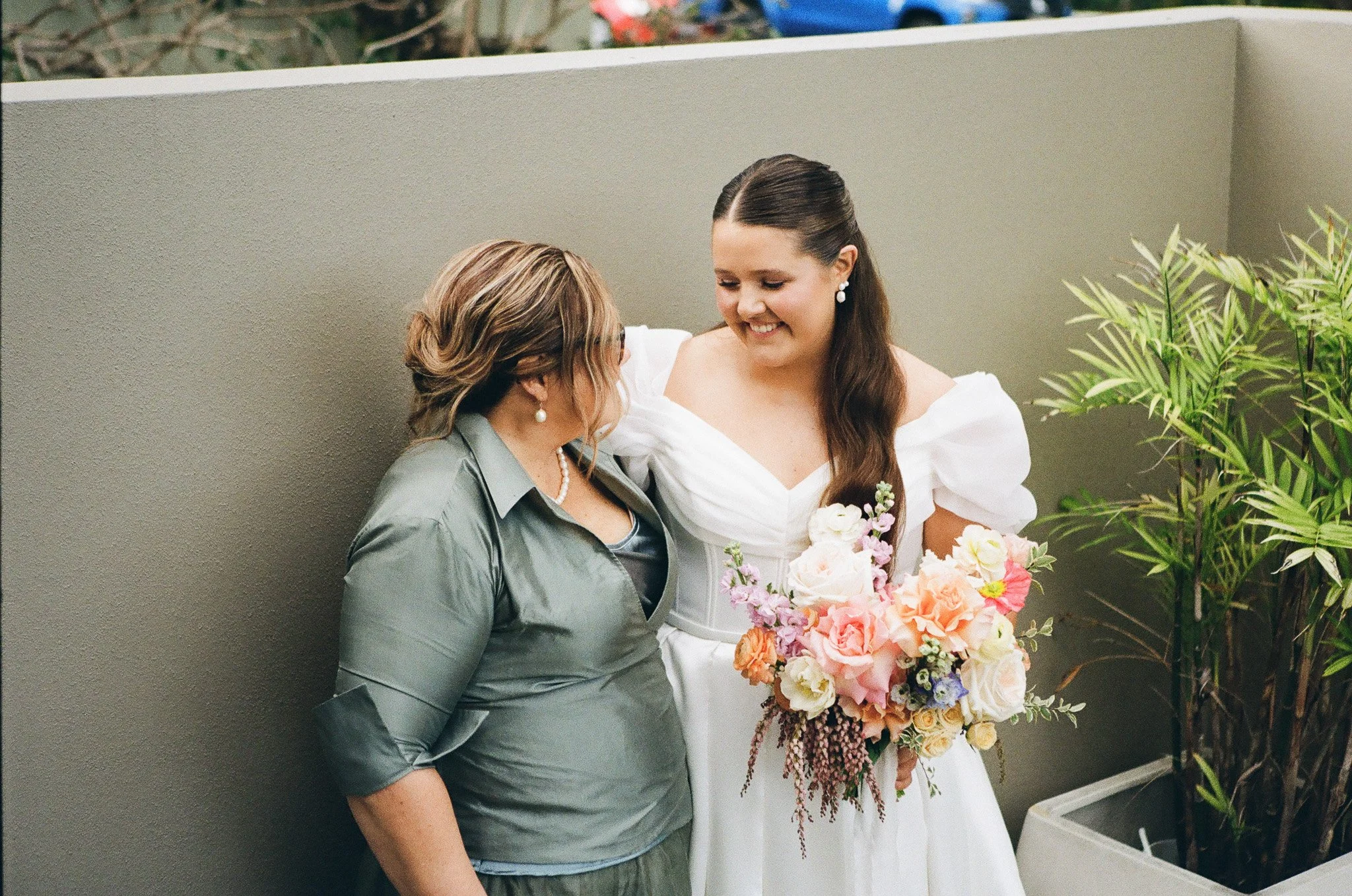 A bride in a white dress holding a colorful bouquet standing next to a woman in a gray top with pearl jewelry, both smiling and looking at each other near a gray wall and a green plant.