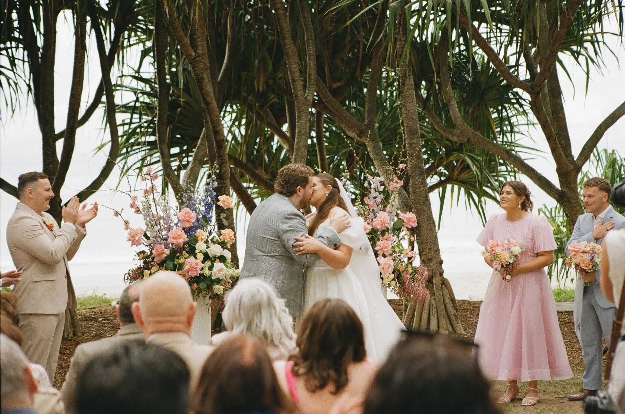 A wedding ceremony on the beach with the bride and groom kissing under trees, surrounded by floral arrangements and guests applauding and smiling.