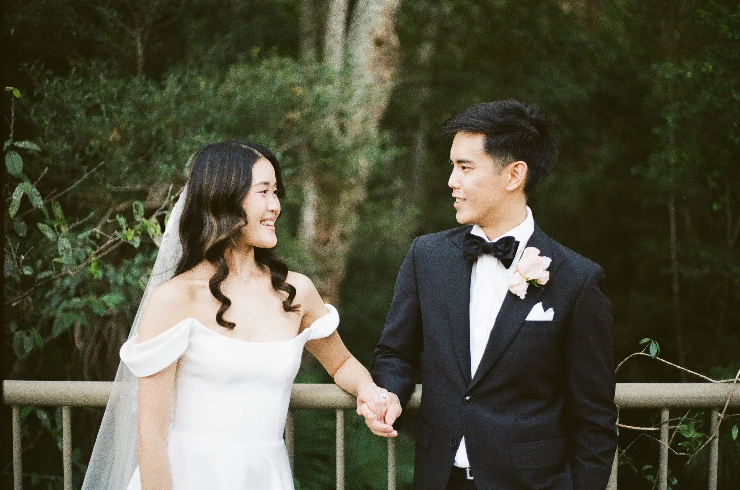 A bride and groom holding hands and smiling at each other outdoors, with trees in the background.