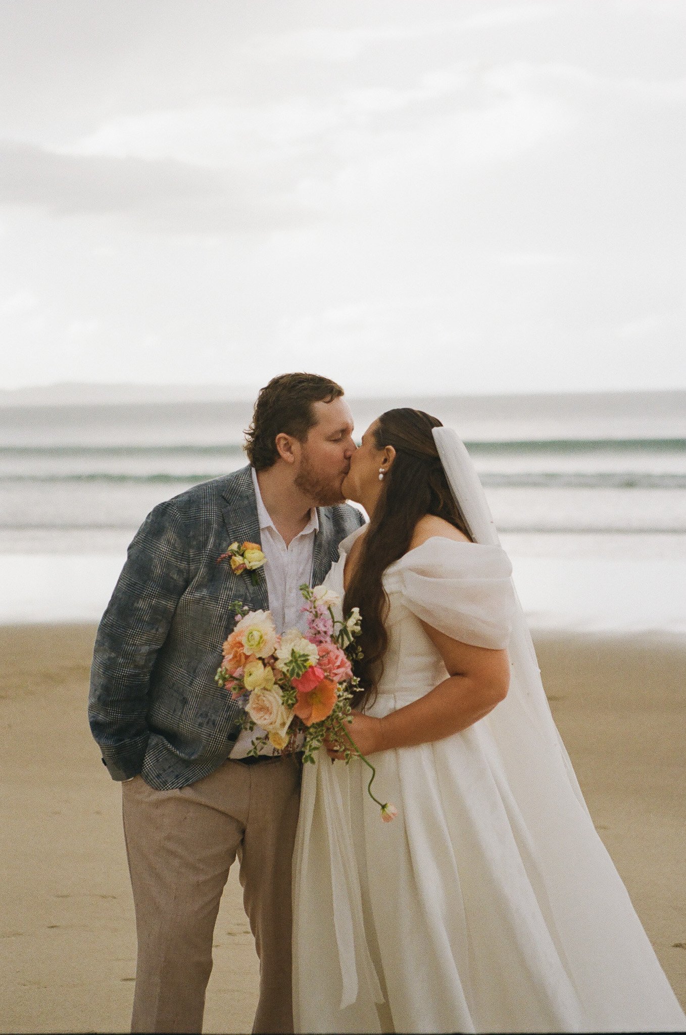 A bride and groom kiss on a beach during their wedding, with the ocean and cloudy sky in the background. The bride is holding a colorful bouquet of flowers.