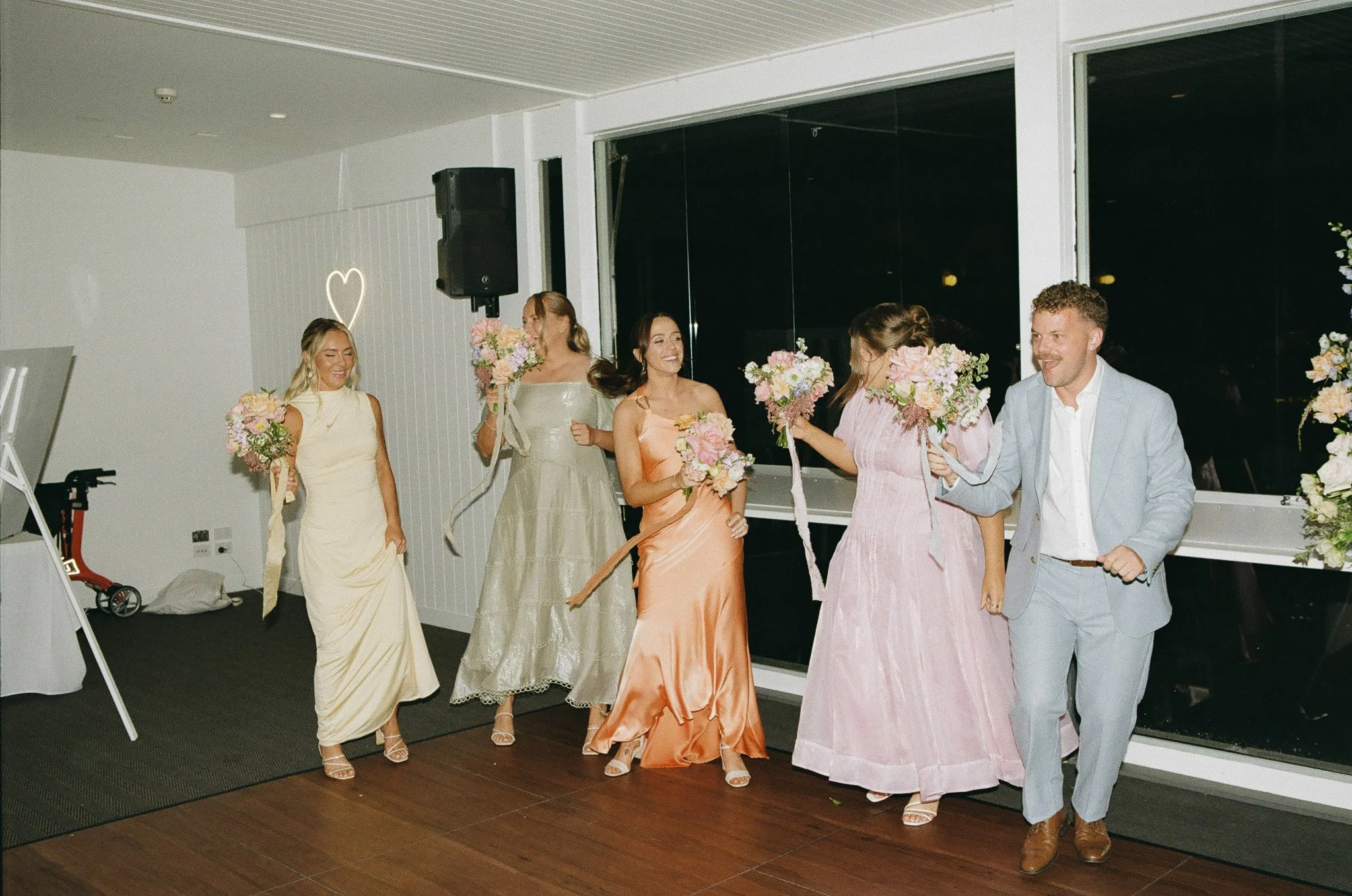 Group of women and one man at a wedding reception, holding bouquets of flowers, dancing or walking across a wooden floor with a large window behind them, decorated with floral arrangements.
