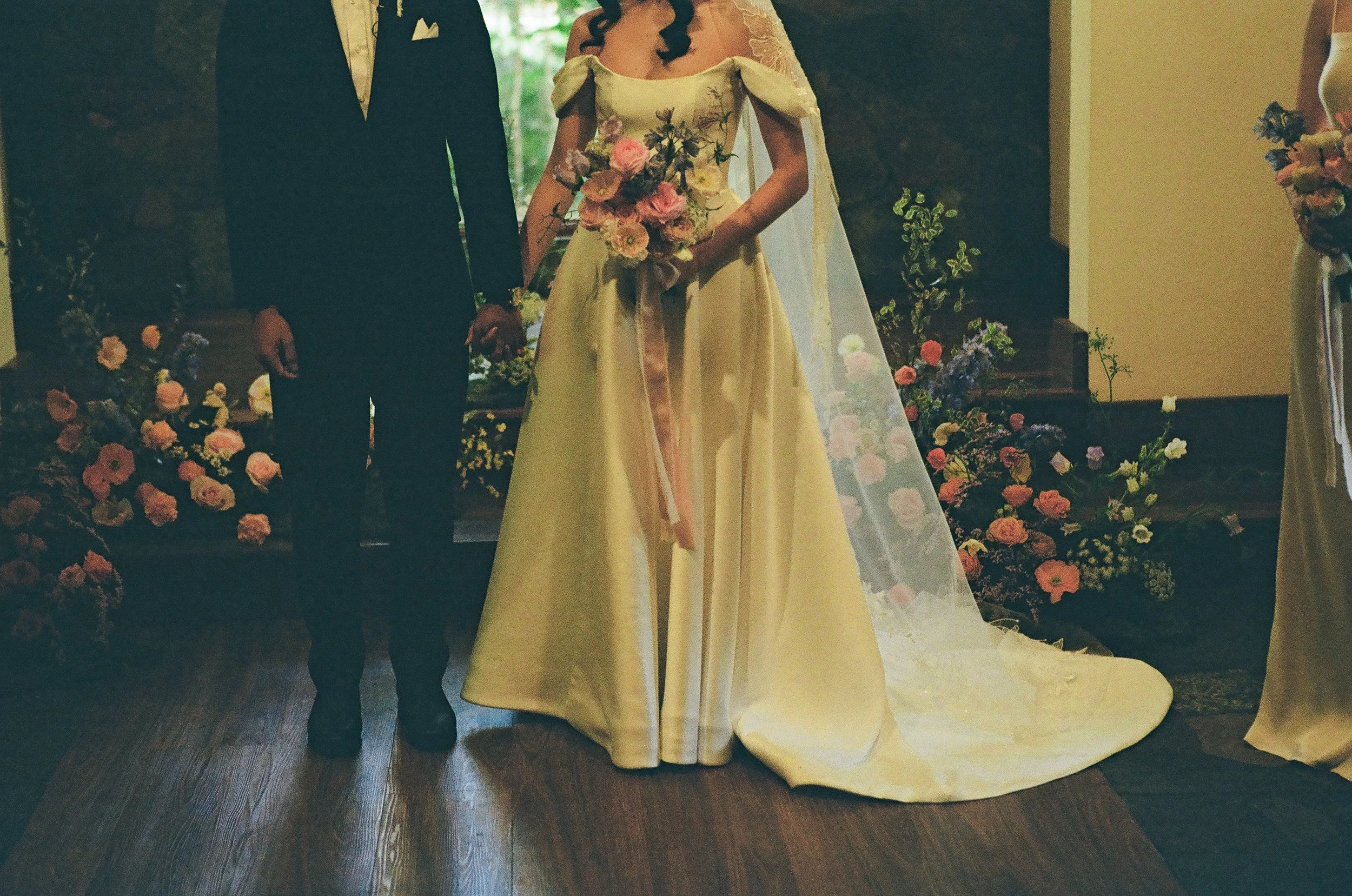 A bride and groom standing together during a wedding ceremony, holding hands. The bride wears a yellow gown with floral details and a veil, holding a bouquet of pink and purple flowers. The groom wears a black suit with a white shirt. The background 
