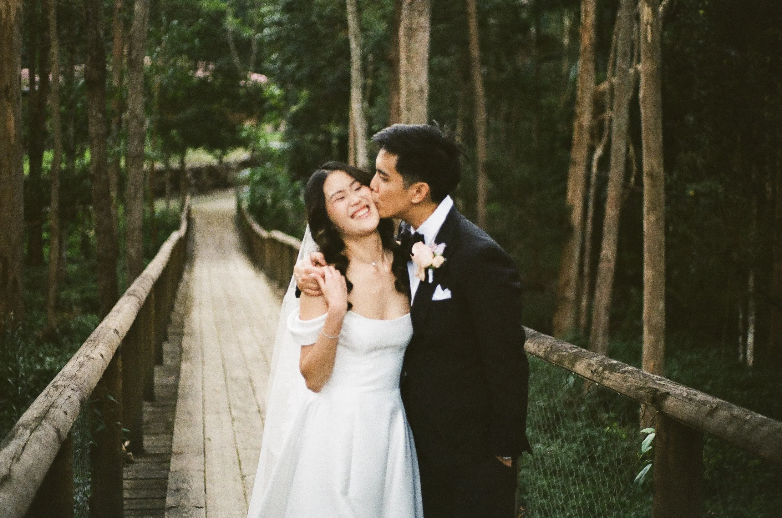 A wedding couple sharing a kiss on a wooden bridge in a forested area, with the bride in a white dress and veil, and the groom in a black tuxedo with a boutonniere.