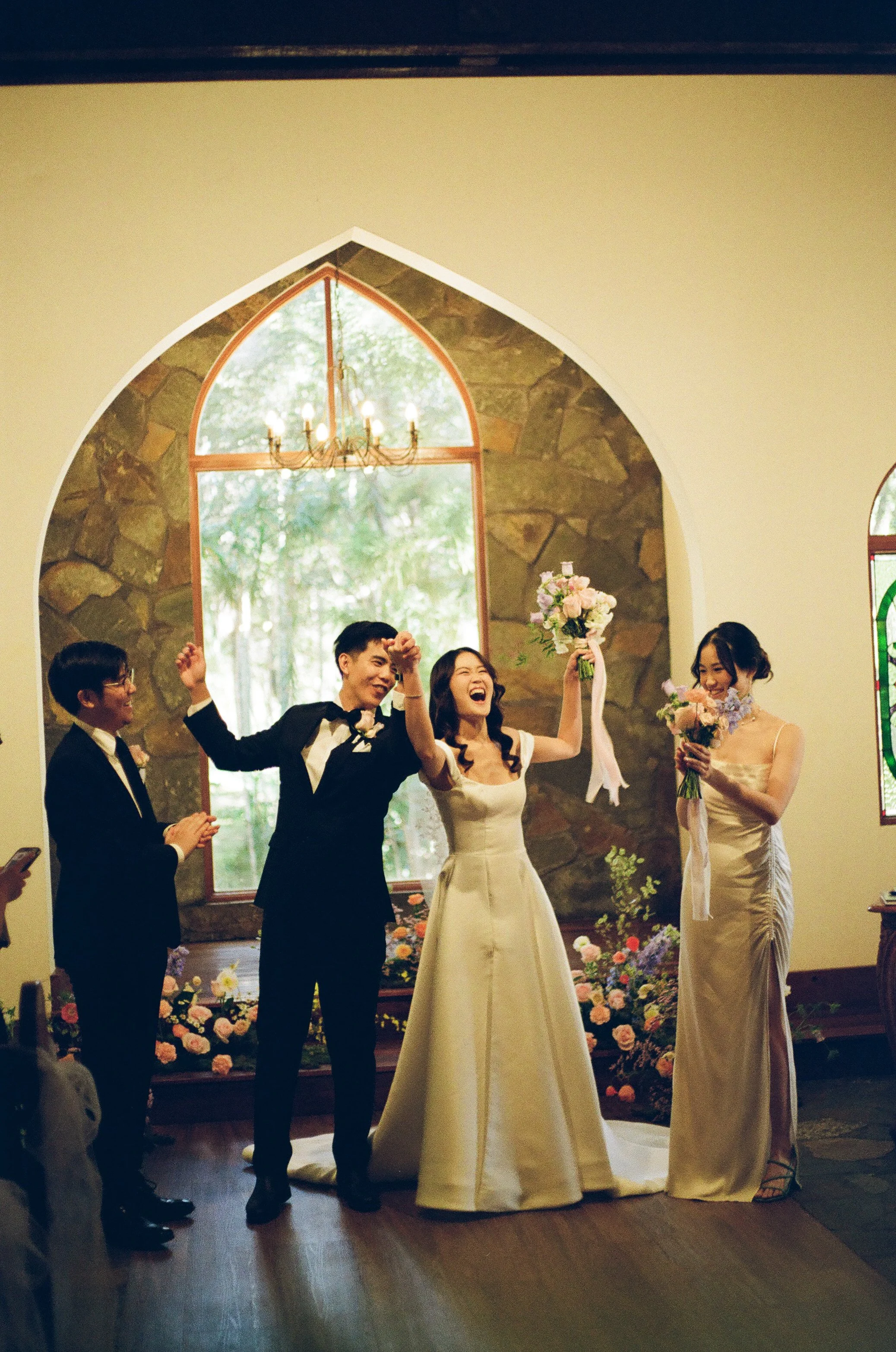 A wedding ceremony inside a church with a large arched window in the background. The bride, dressed in a white gown, is holding a bouquet and appears happy. The groom, in a black tuxedo, is holding her hand and smiling. Two women beside them are also holding bouquets, dressed in matching beige dresses, and smiling. There are floral decorations on the floor in front of the window.