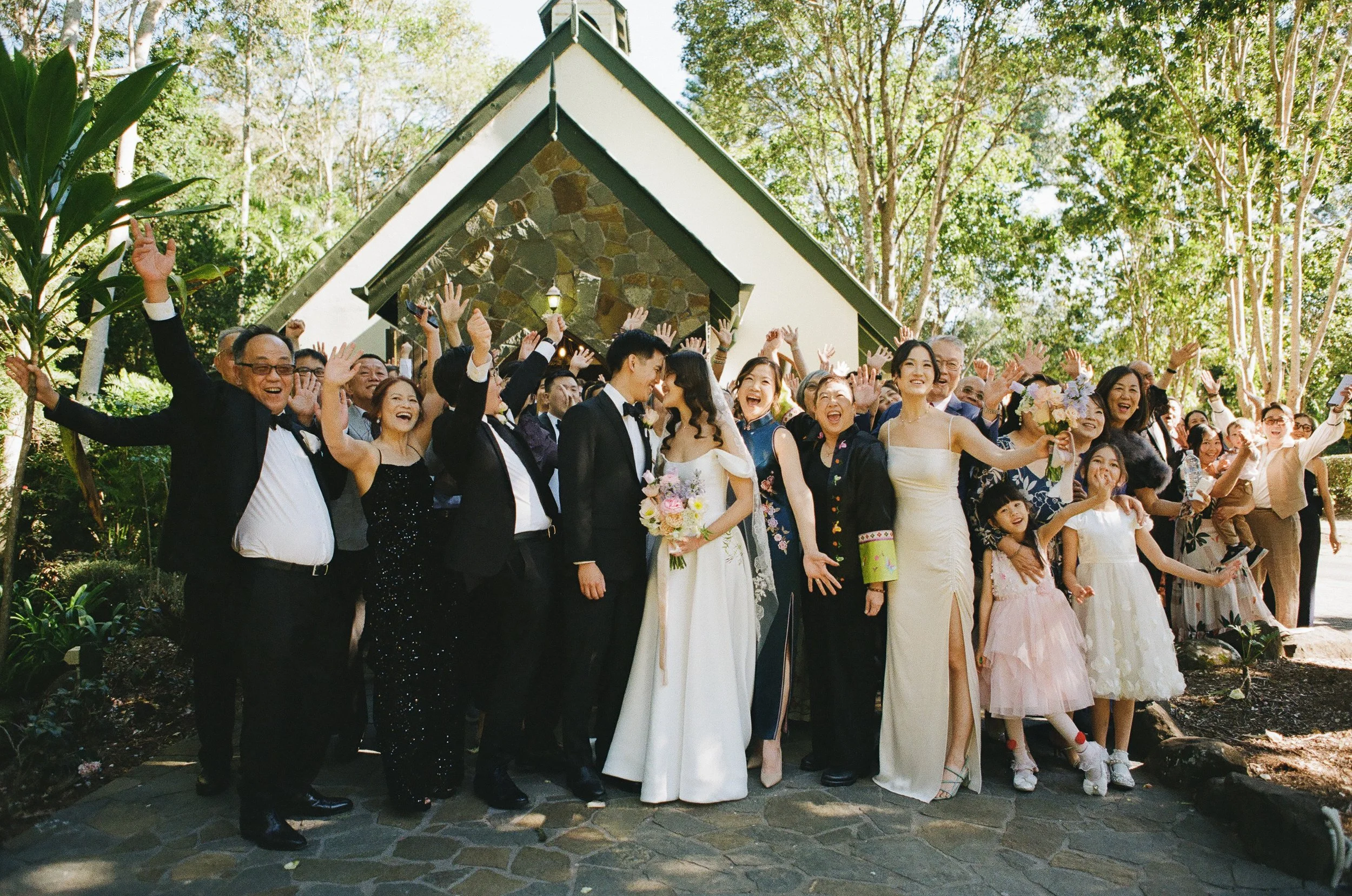 A large group of people at a wedding celebration outside, dressed in formal attire, smiling and waving, with the bride and groom in the center, surrounded by family and friends.