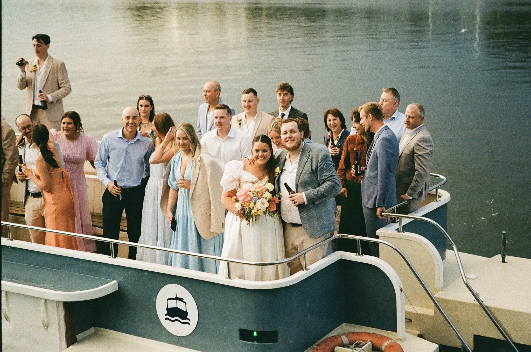 Group of wedding guests on a boat by the water, celebrating and smiling, with some holding drinks, the bride holding a bouquet.