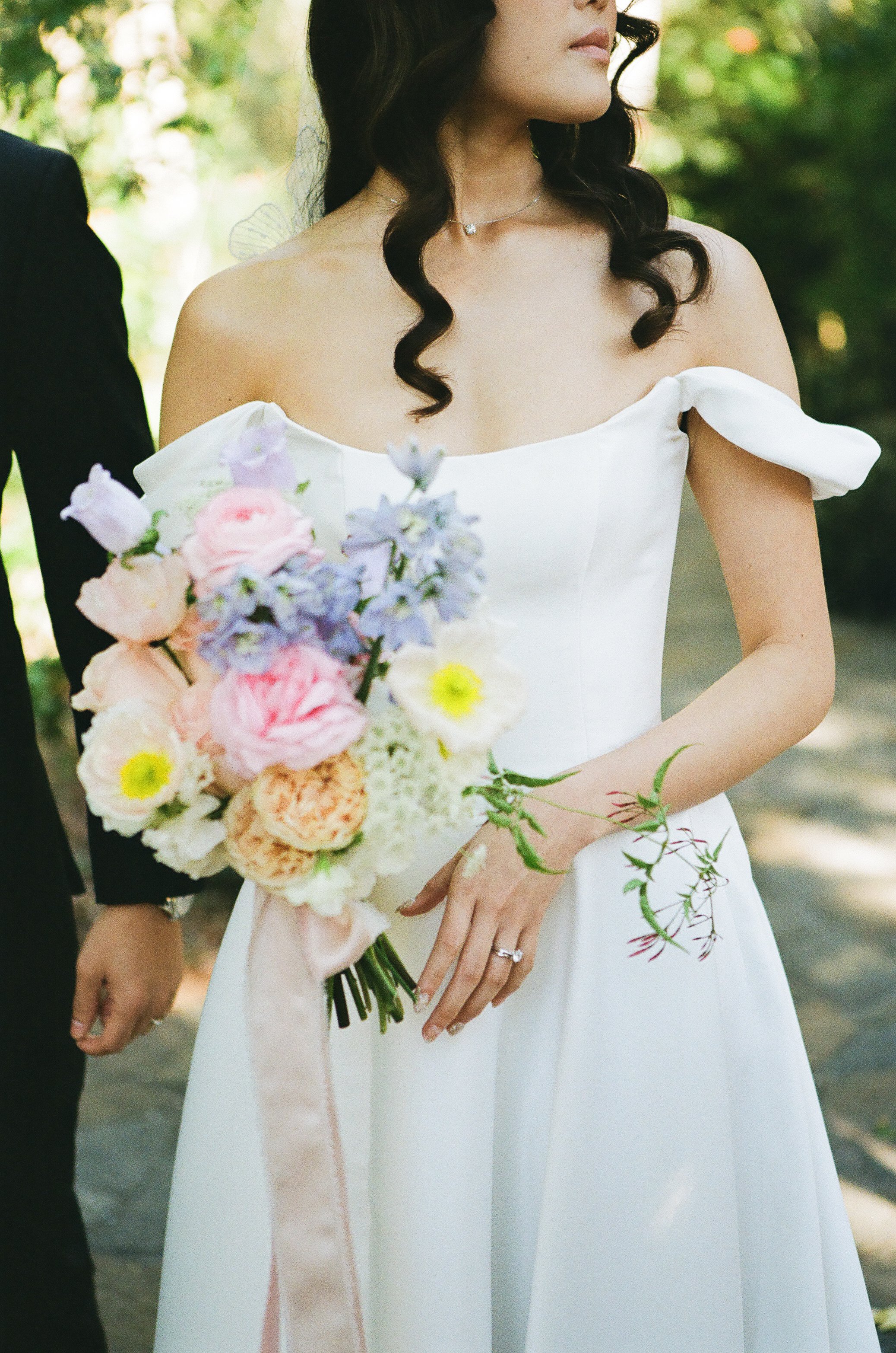 A bride holding a bouquet of pastel pink, peach, purple, and yellow flowers during a wedding ceremony outdoors.