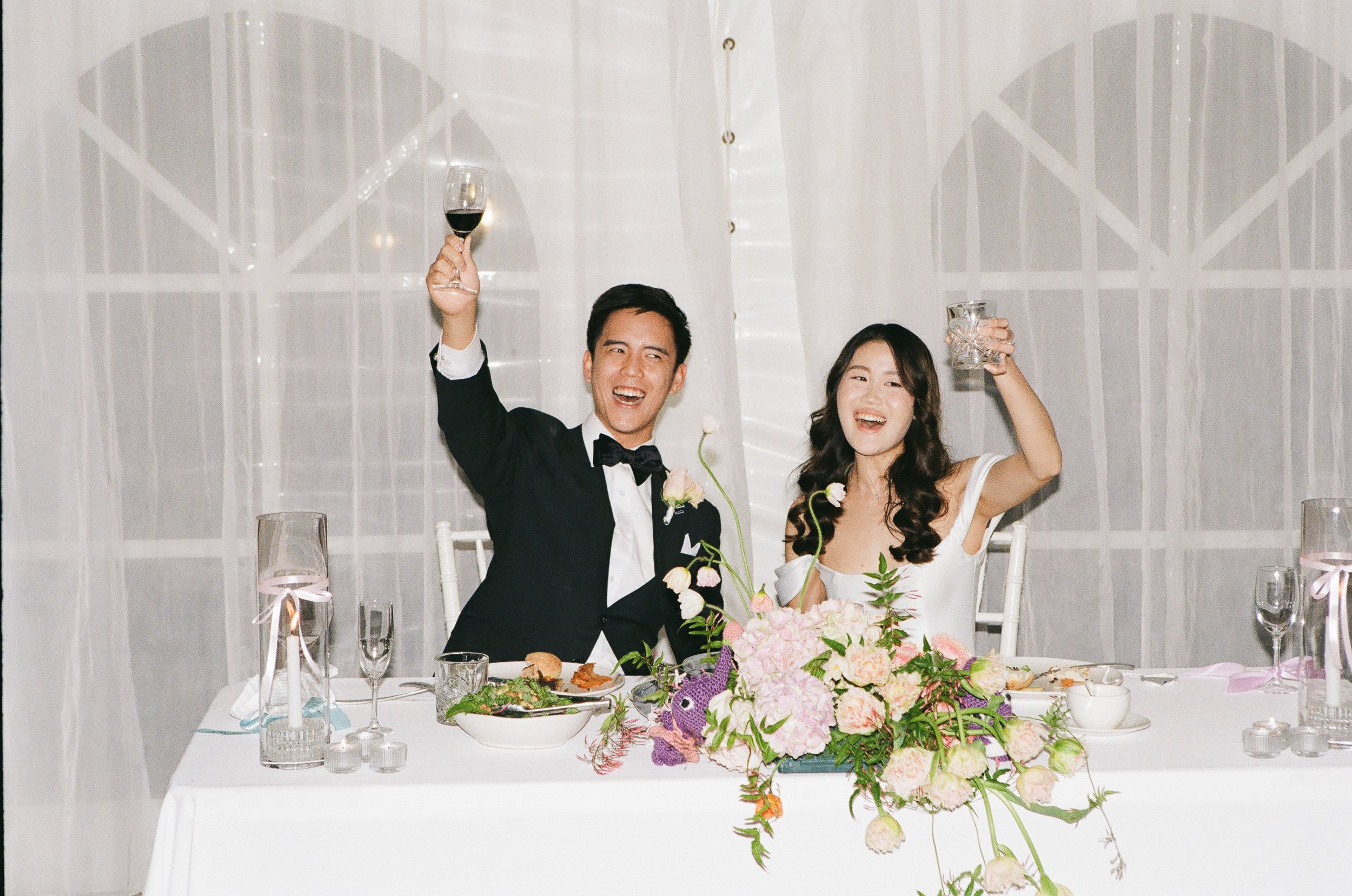 A newly married couple dressed in wedding attire celebrating with drinks at their wedding reception. The groom is in a tuxedo and the bride in a white dress, seated at a decorated table with flowers and food.