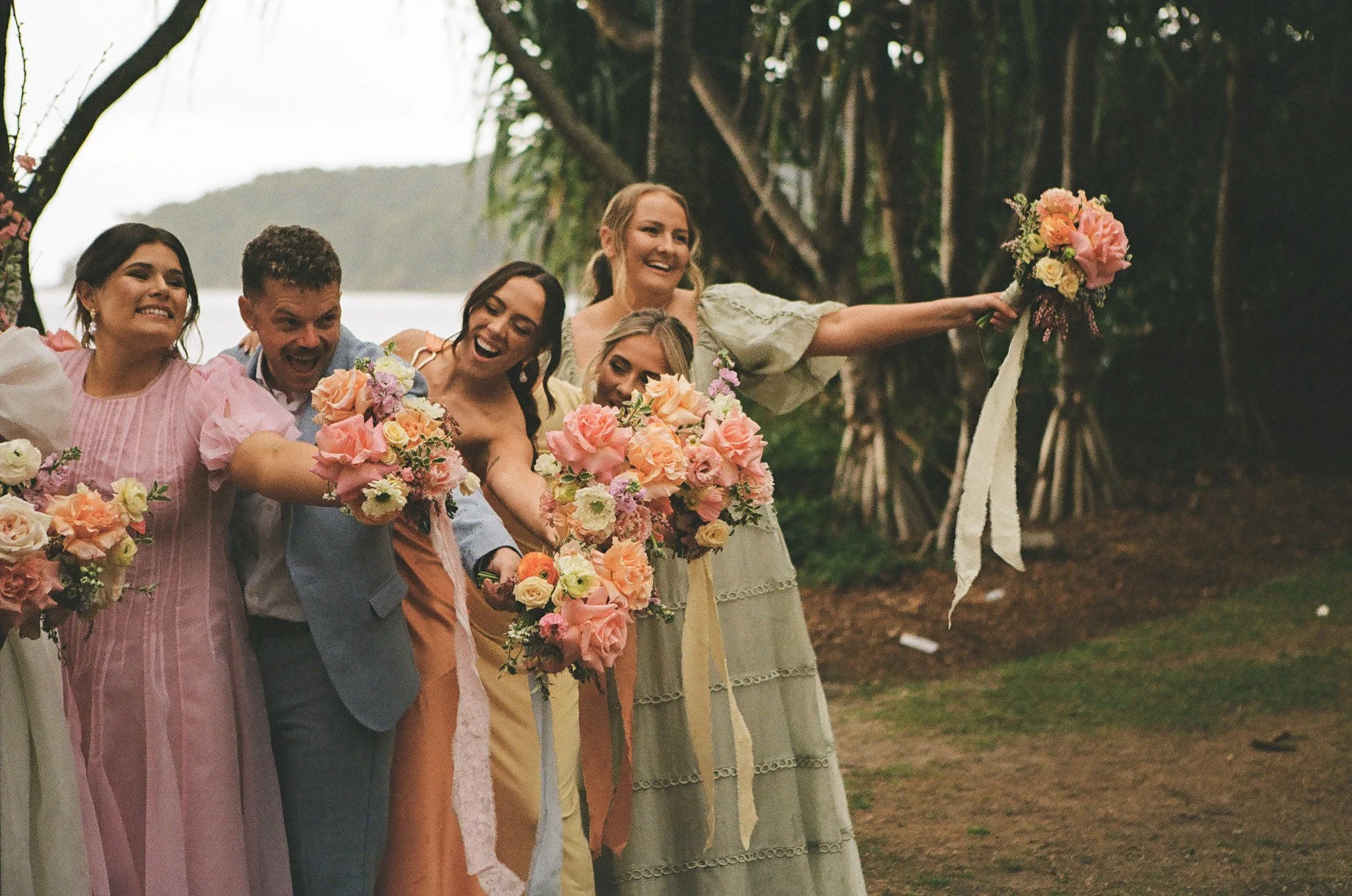 A group of people, including women and a man, smiling and holding bouquets of flowers at an outdoor wedding or celebration, with trees and a body of water in the background.