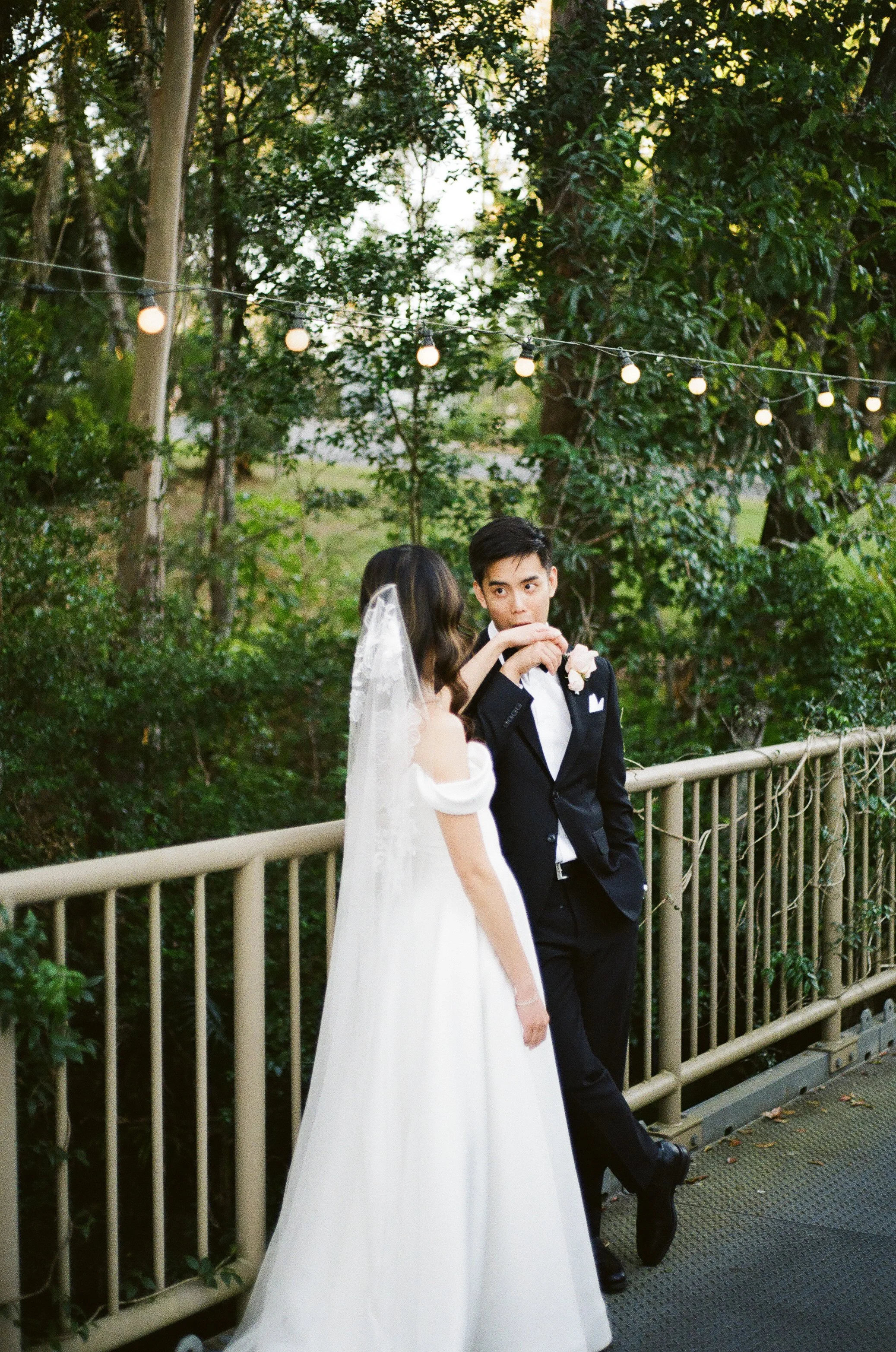 A bride in a white wedding dress and veil, and a groom in a black tuxedo, standing together outdoors near a wooden railing with string lights overhead, surrounded by trees and greenery.