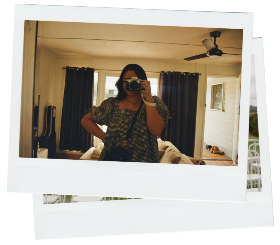 Woman taking a mirror selfie with a camera in a living room with dark curtains, a ceiling fan, and a white house exterior visible through the window.