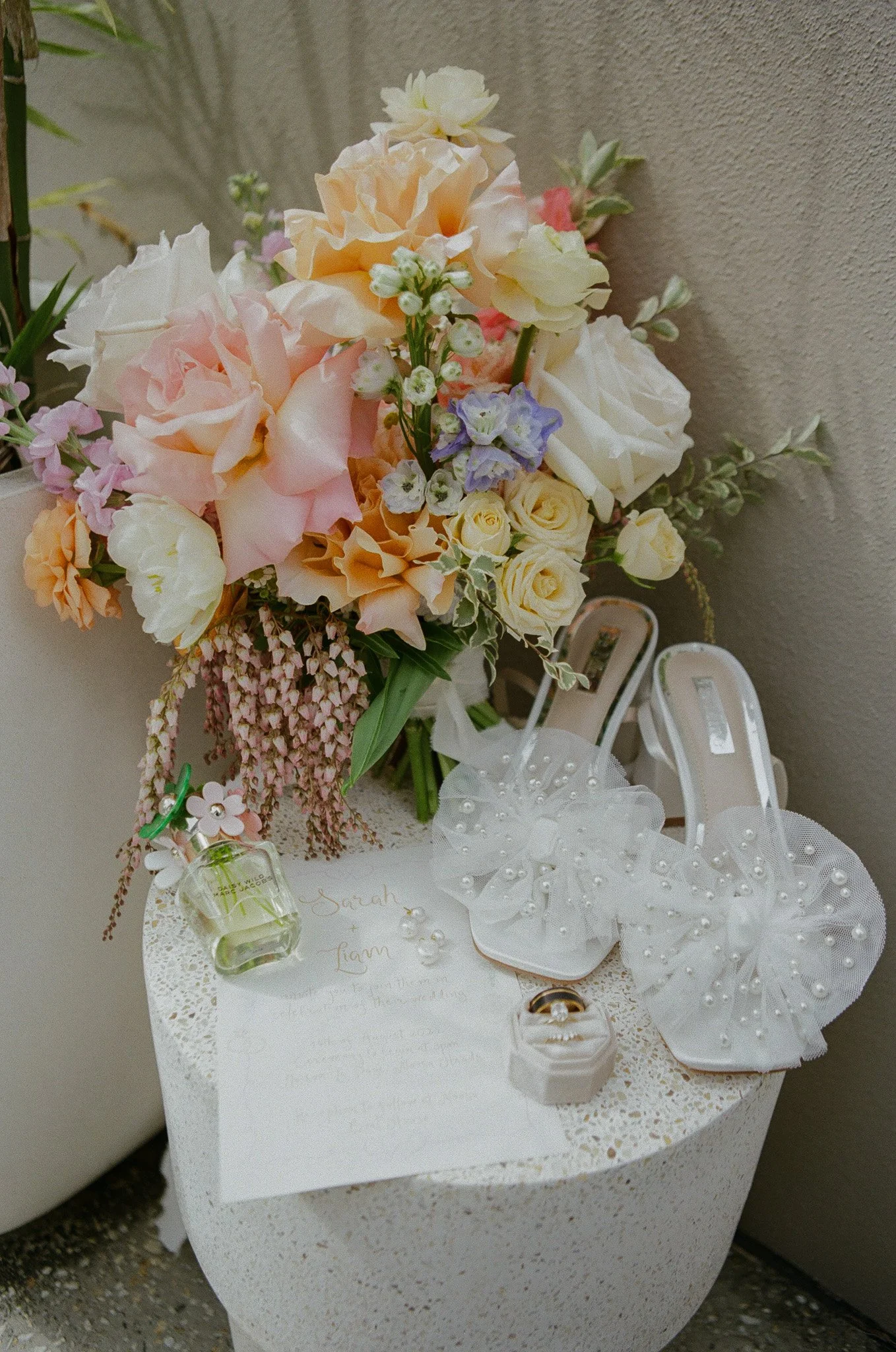 A floral arrangement with pastel roses and other flowers, wedding rings in a white box, a handwritten note, and decorative shoes with pearls on a round white table