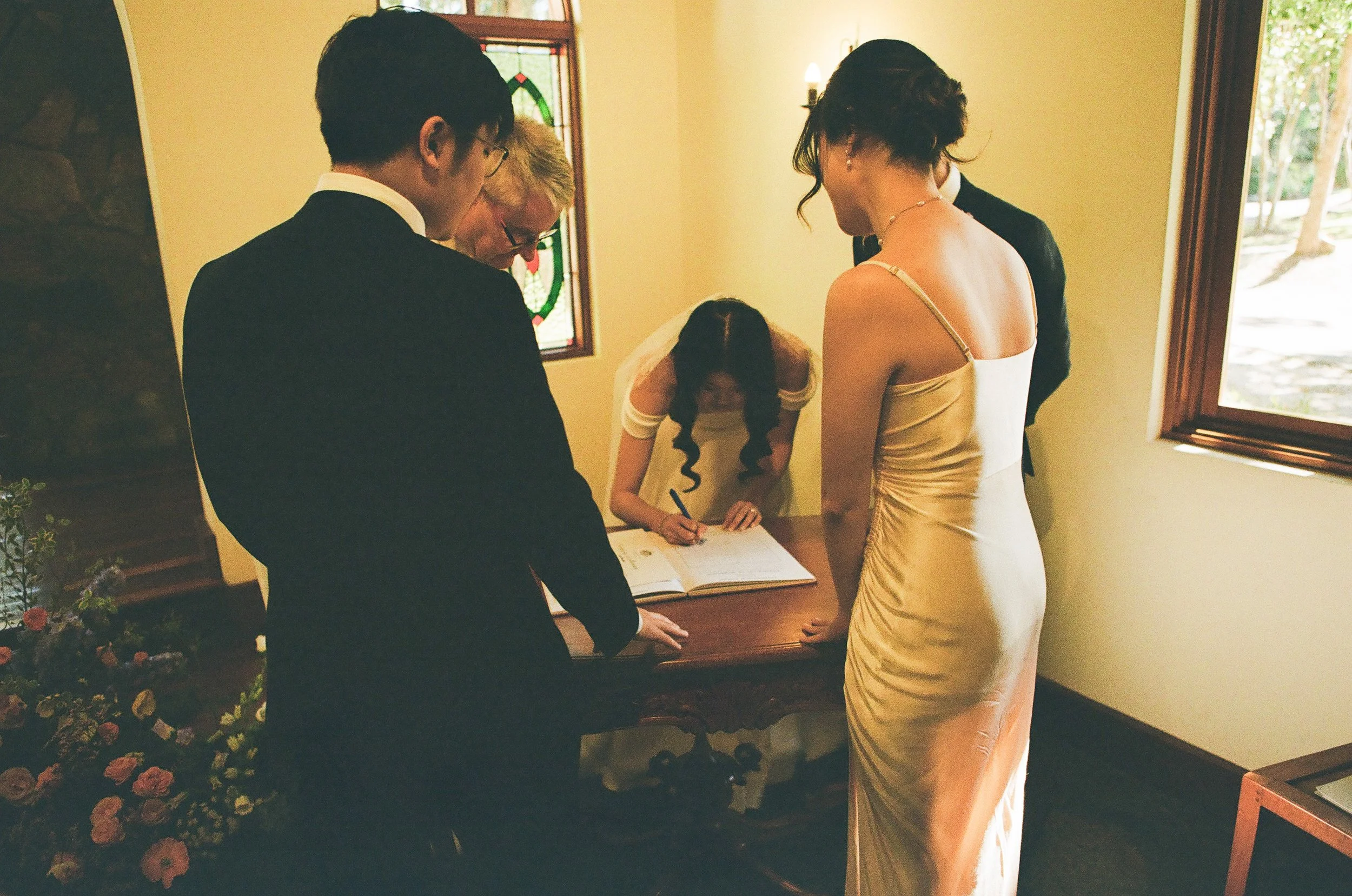 A wedding ceremony with a bride signing a marriage register at a small table, surrounded by guests, inside a room with stained glass windows and large windows allowing natural light.