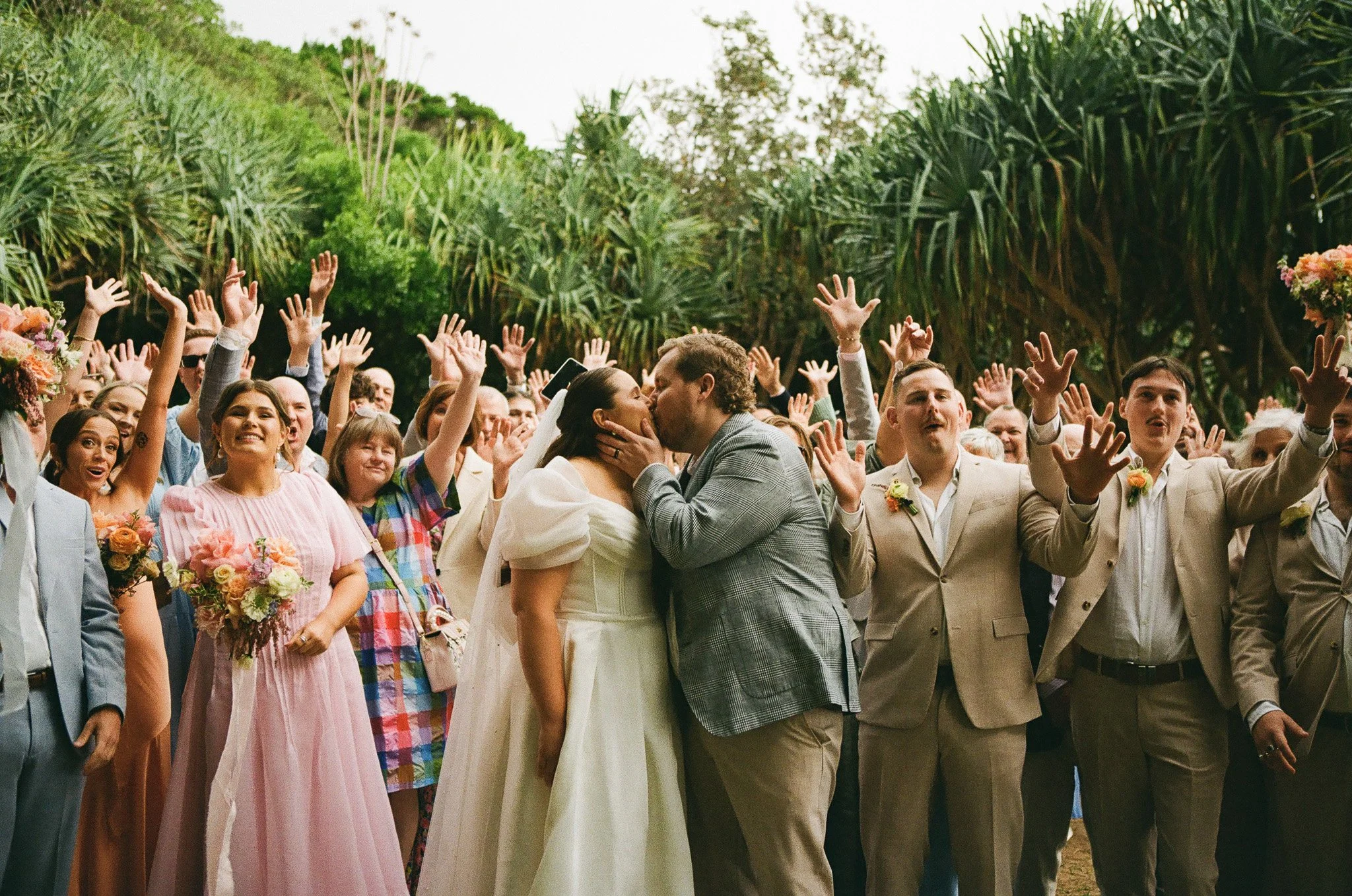 A wedding celebration outdoors with the bride and groom kissing in the center amidst a crowd of wedding guests raising their hands.