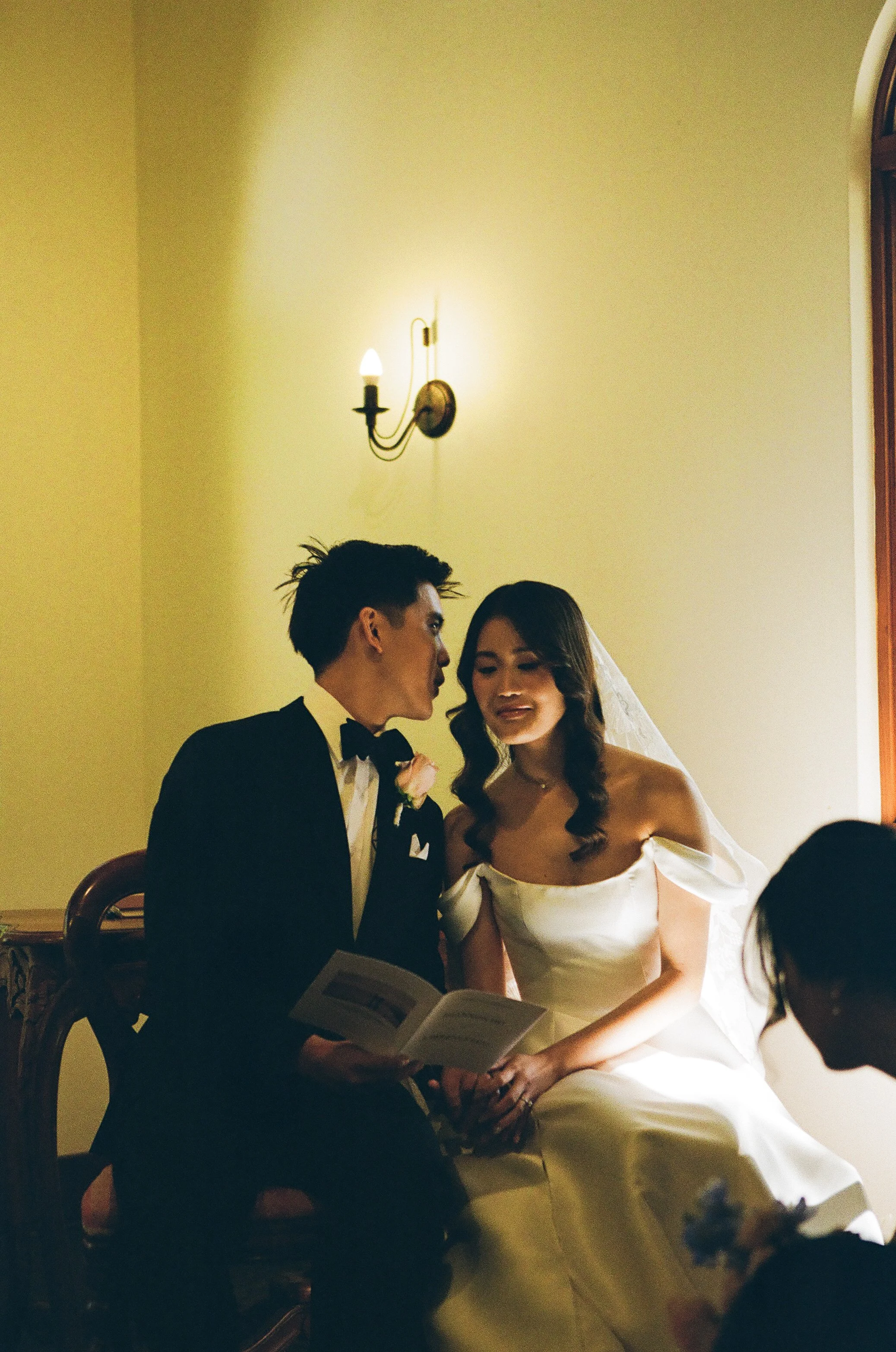 A bride and groom sitting on a bench during their wedding ceremony, reading vows, with a minimalistic wall light above them.