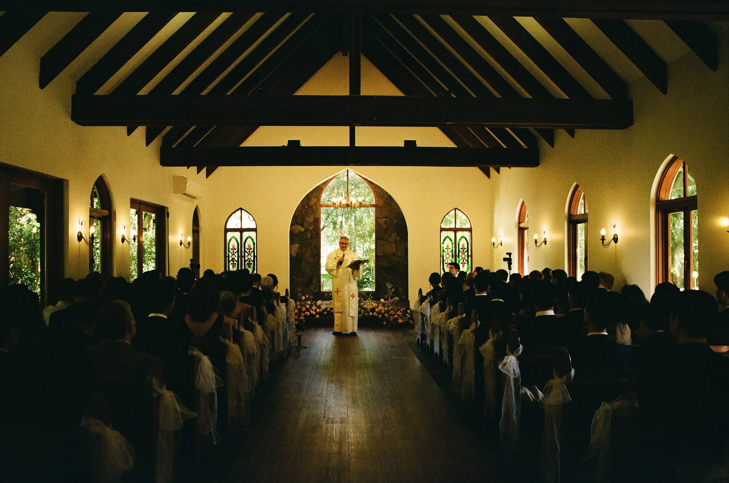 A religious ceremony inside a church with a priest at the altar, surrounded by seated attendees. The church has stained glass windows, wooden beams on the ceiling, and soft lighting.