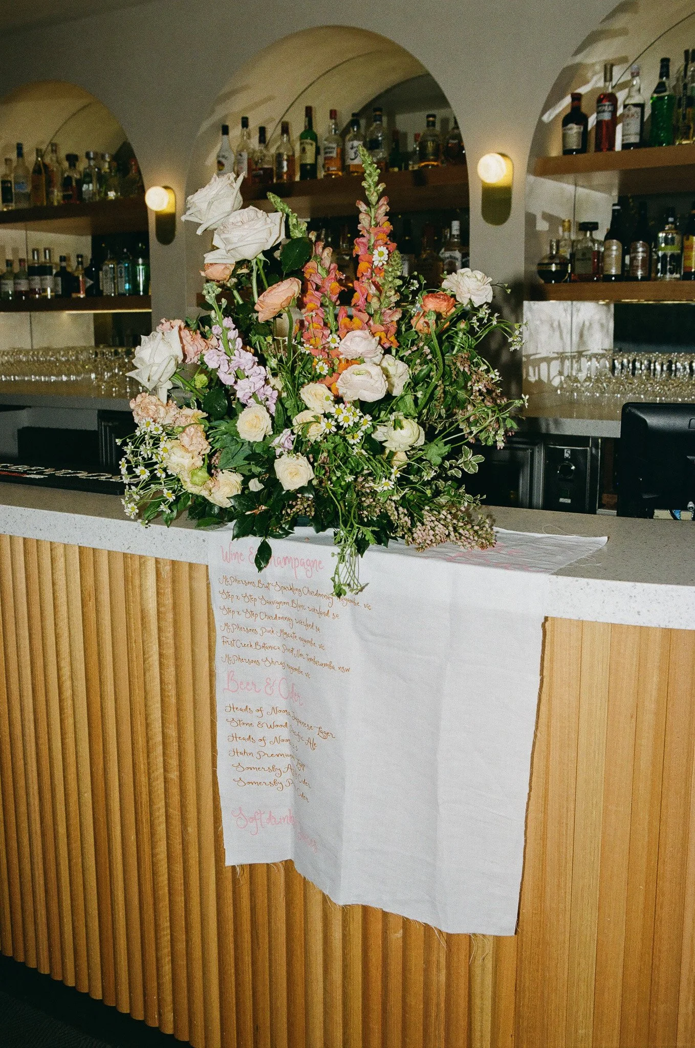 A large floral arrangement with pink, white, and peach flowers on a bar counter, with a menu card hanging underneath.