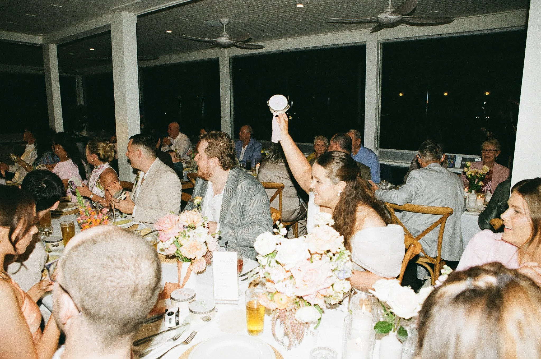 People dining at a formal event with floral centerpieces, some are engaged with their phones, and one woman is raising her hand holding a small mirror.