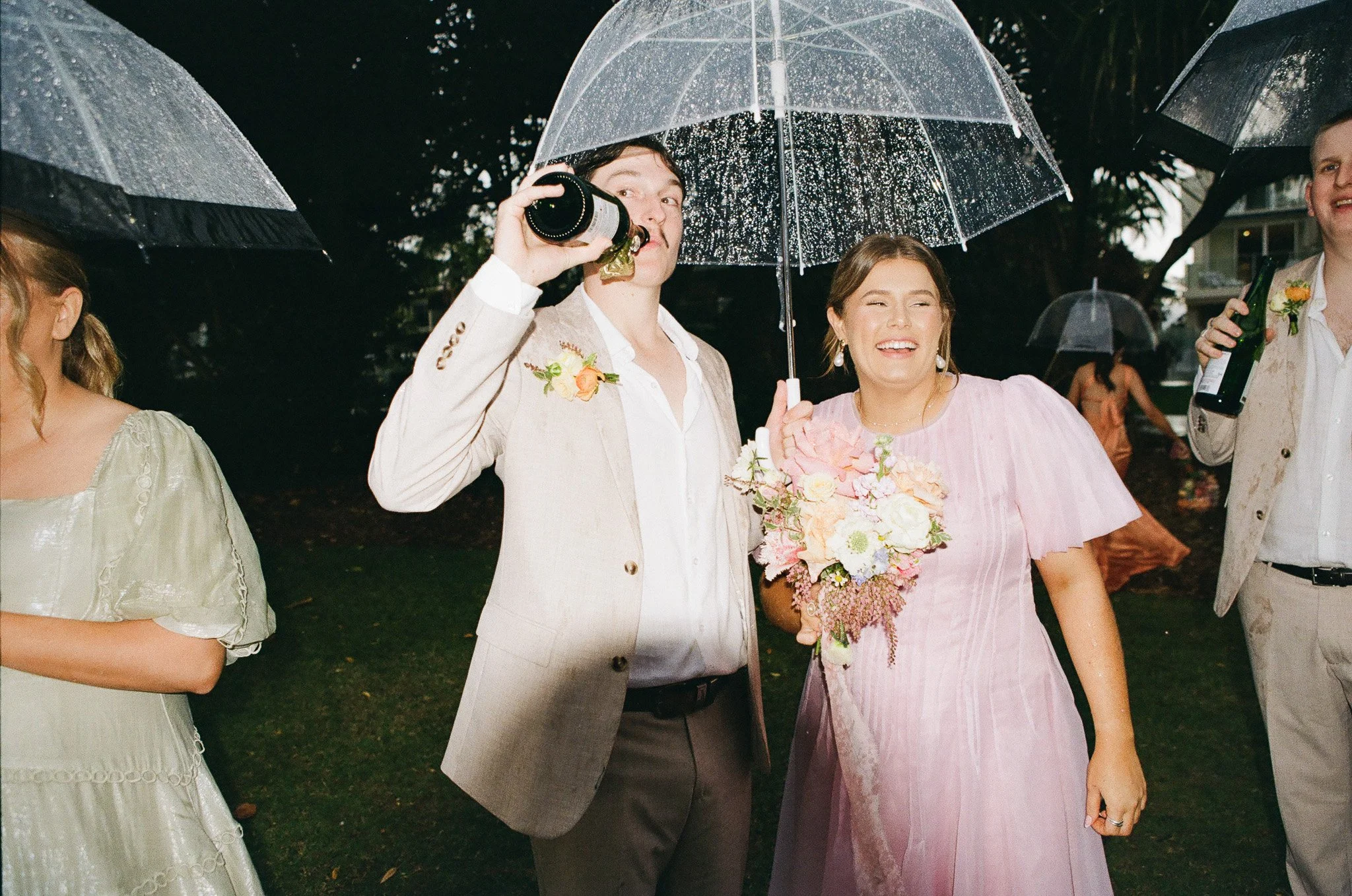 A group of people at a wedding celebration standing outdoors in the rain, holding umbrellas. The woman in a pink dress holds a bouquet of flowers, smiling, while the man in a light-colored suit drinks from a bottle.