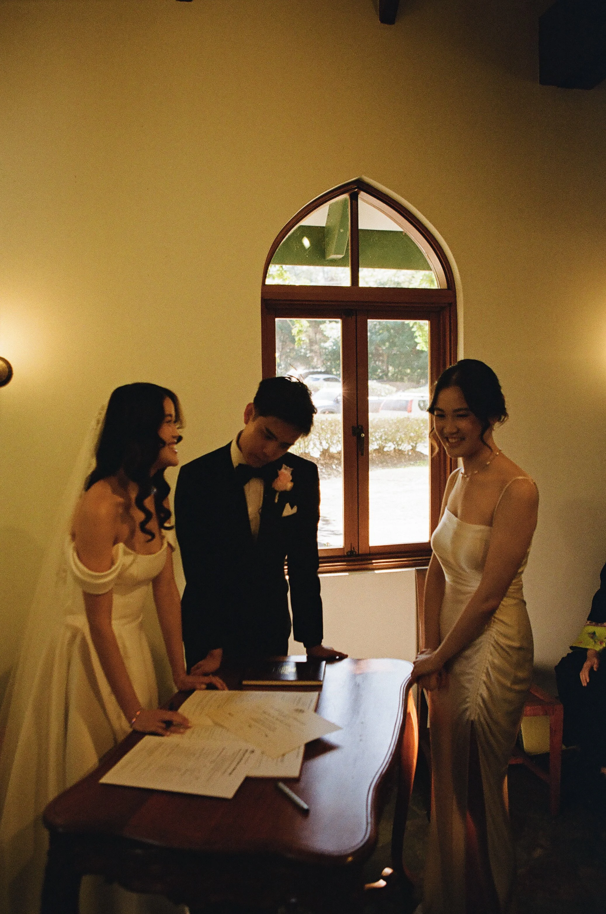 A couple getting married, signing documents with two women standing nearby inside a room with a window and sunlight outside.