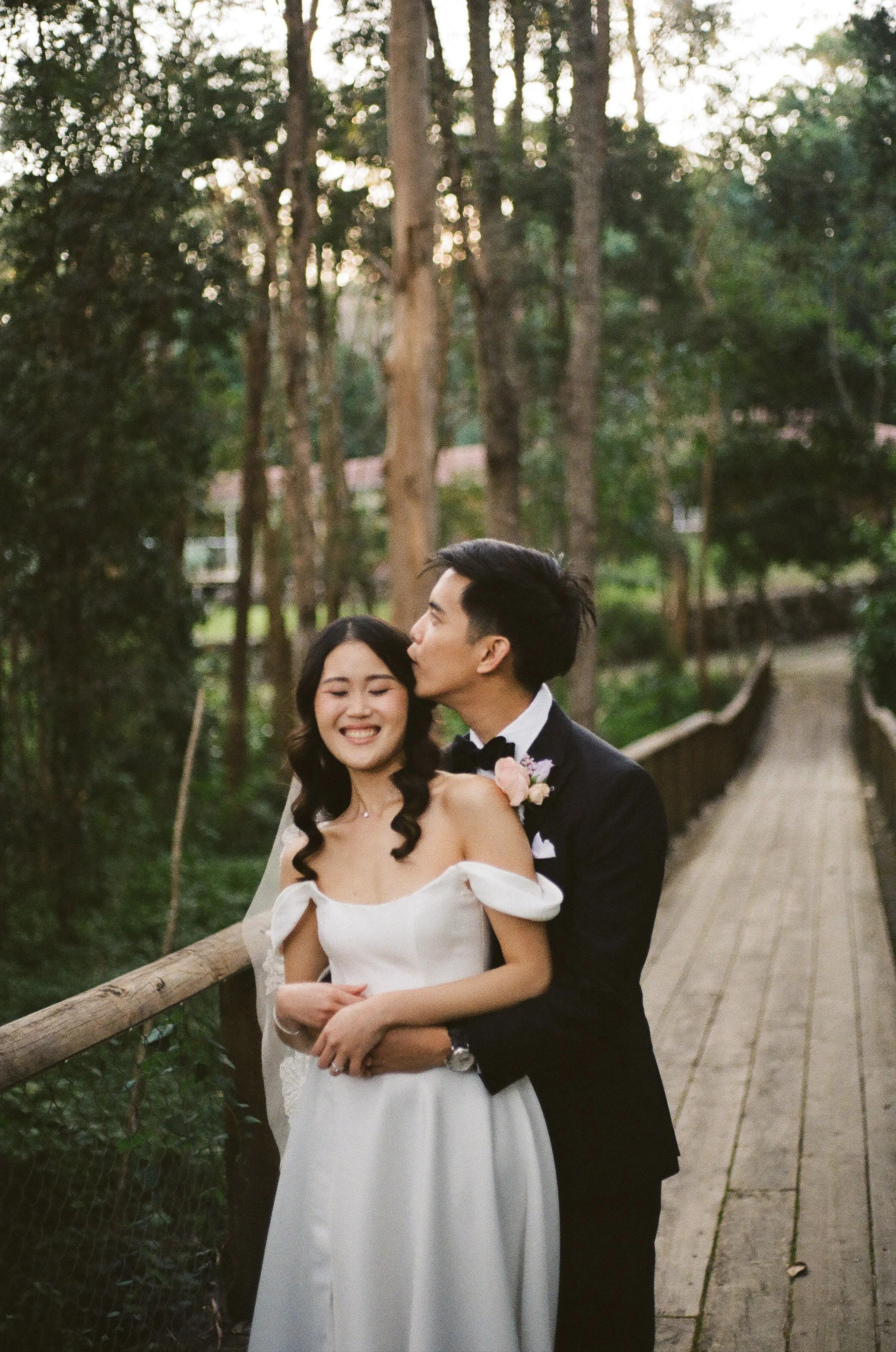 A newlywed couple in wedding attire standing on a wooden bridge in a forest, with the groom kissing the bride on the temple while she smiles with closed eyes.