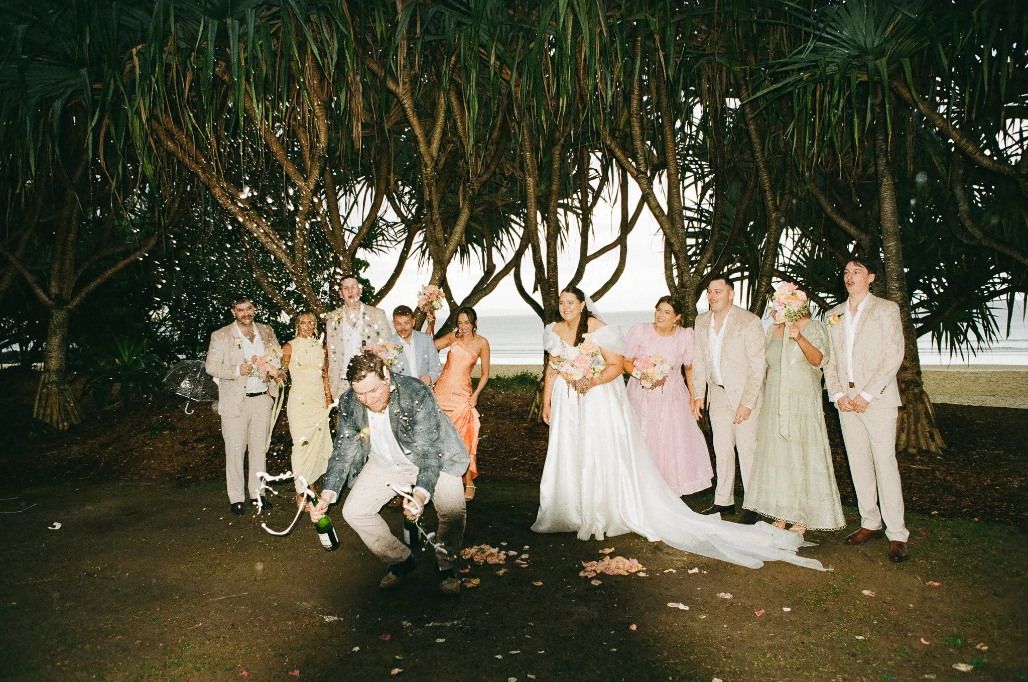 Group of people celebrating at a wedding beneath trees by the beach, with one person opening champagne while others hold bouquets, confetti in the air, and flower petals on the ground.