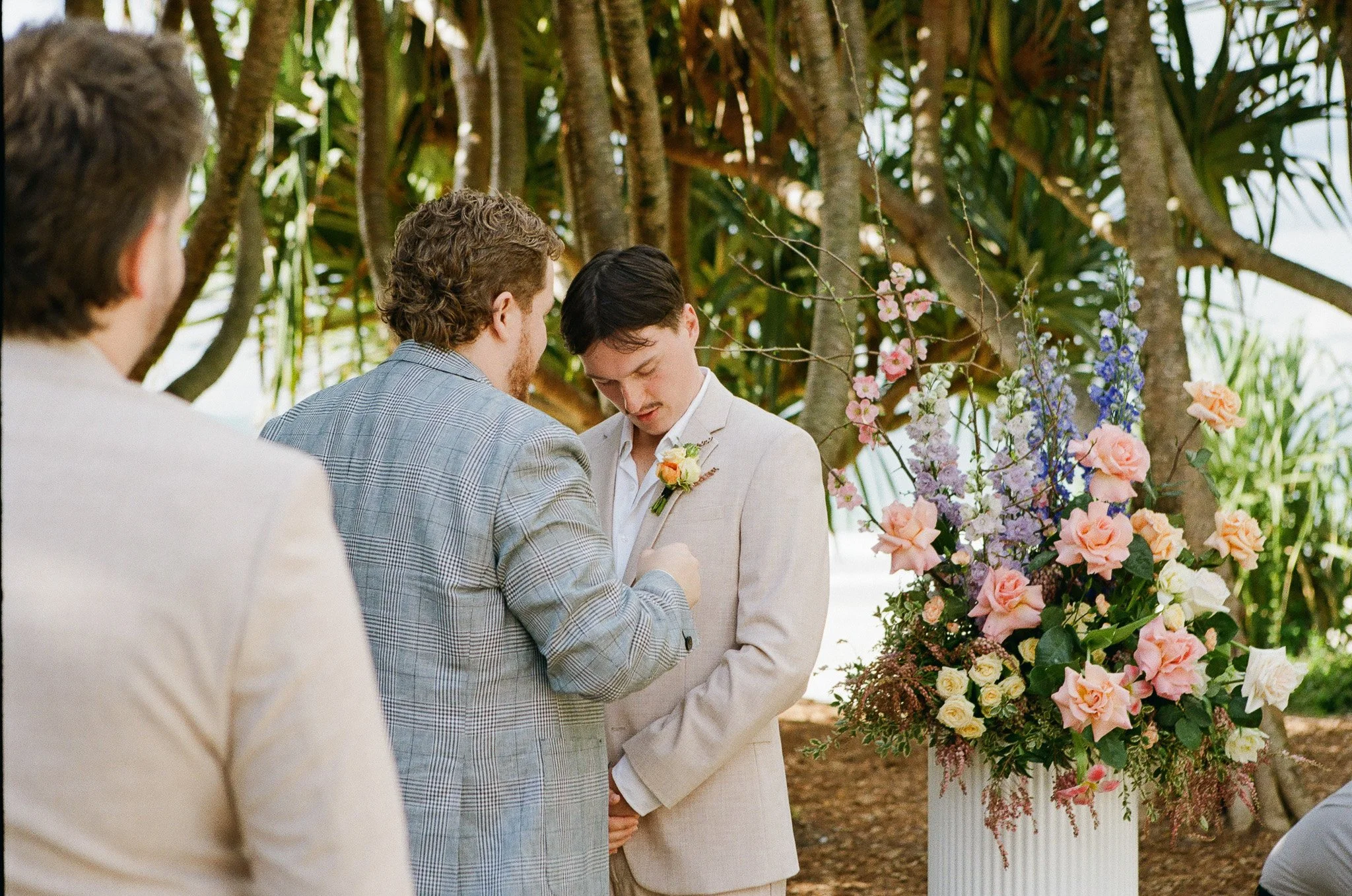 Two men at a wedding altar, one placing a ring on the other's finger, with a large floral arrangement nearby and tropical trees in the background.