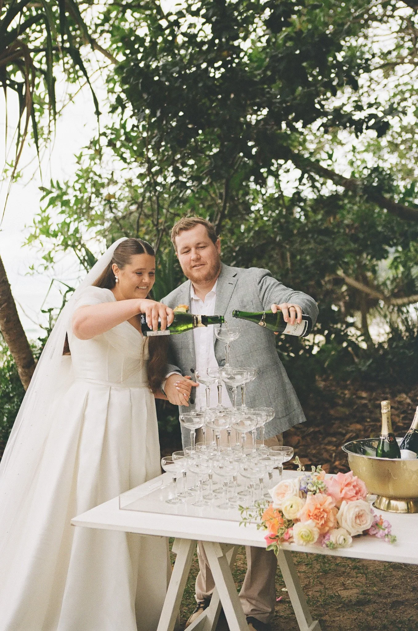 A bride and groom at a wedding celebration are pouring champagne into a pyramid of glasses outdoors, with a table of flowers and bottles nearby.