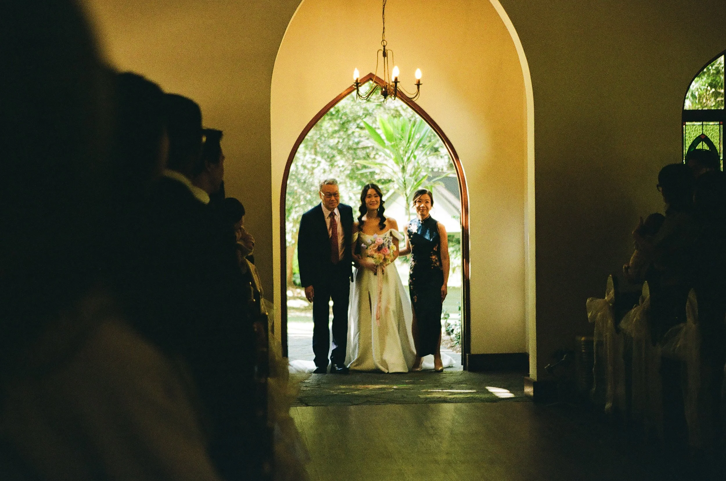 Bride in white dress holding a bouquet of flowers, flanked by two people, walking out of a church with an arched doorway, with guests seated on either side in shadow.