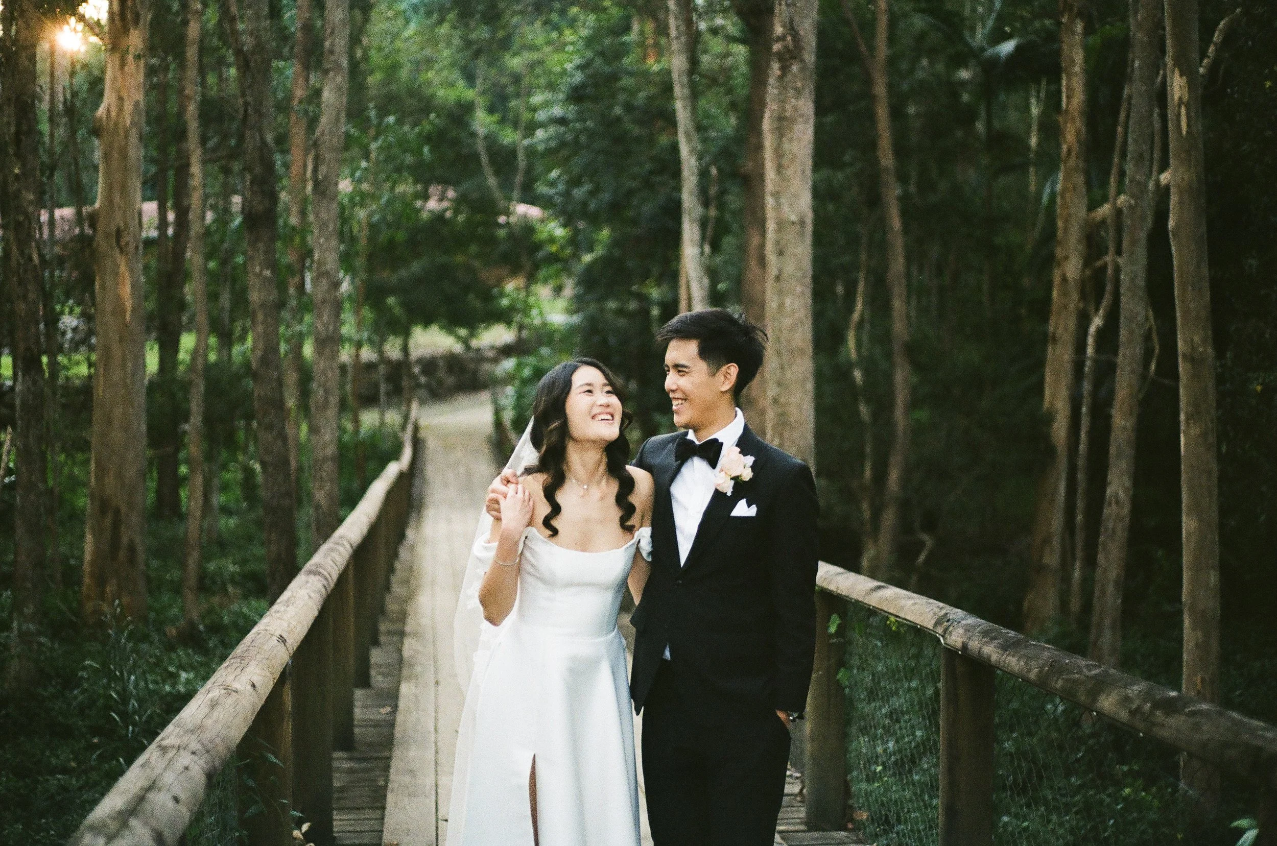 A bride and groom smiling and looking at each other on a wooden bridge surrounded by trees.