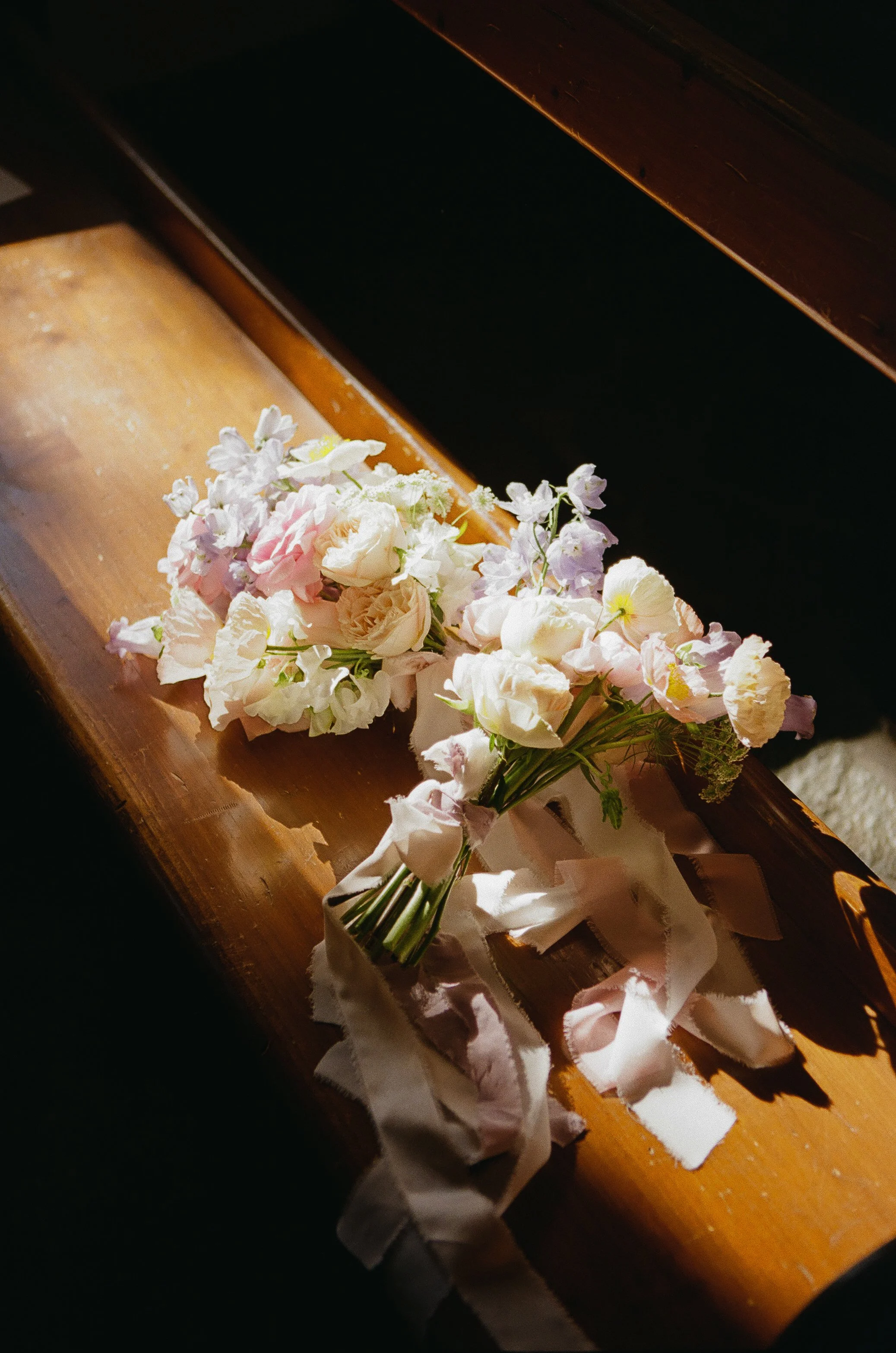 A small bouquet of delicate white, pink, and purple flowers with green stems and a white ribbon on a polished wooden surface, with sunlight casting shadows.