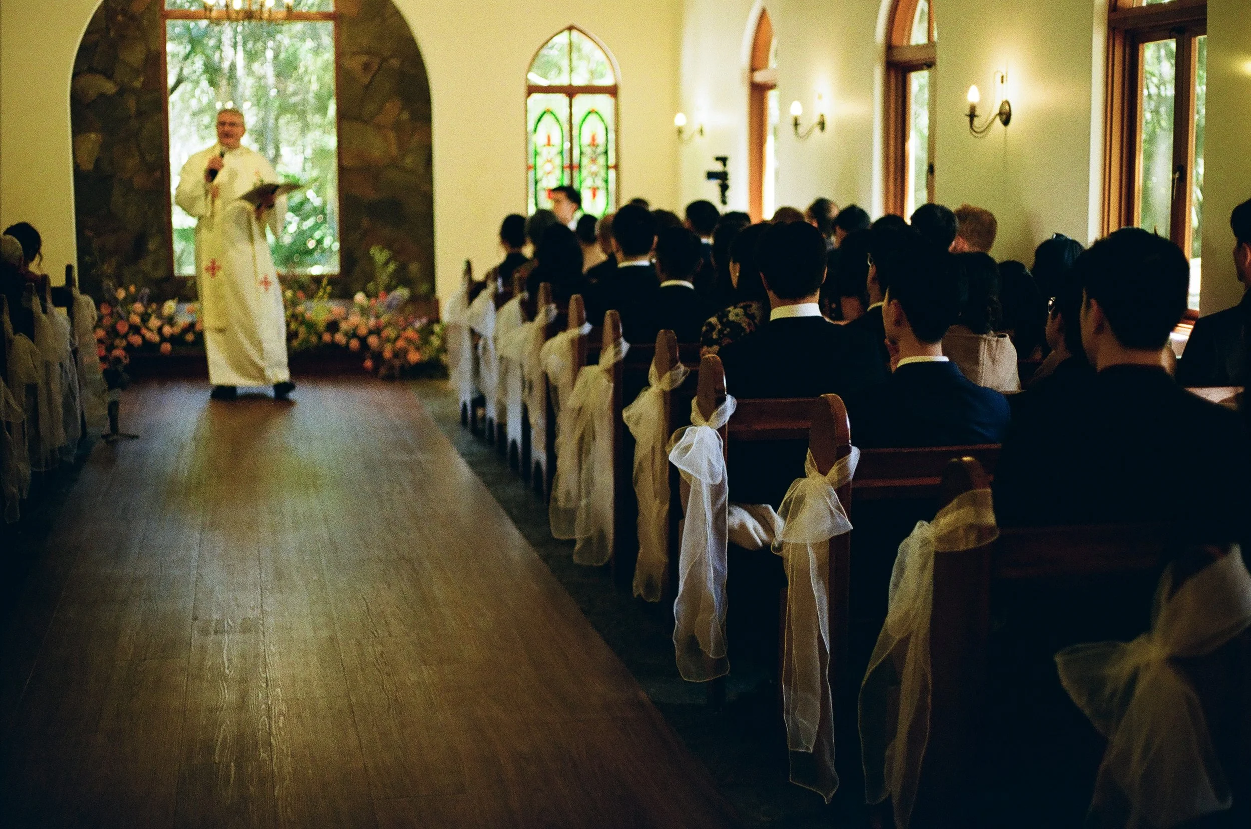 A priest officiates a wedding ceremony in a small chapel with guests seated in pews decorated with white bows, and stained glass windows in the background.