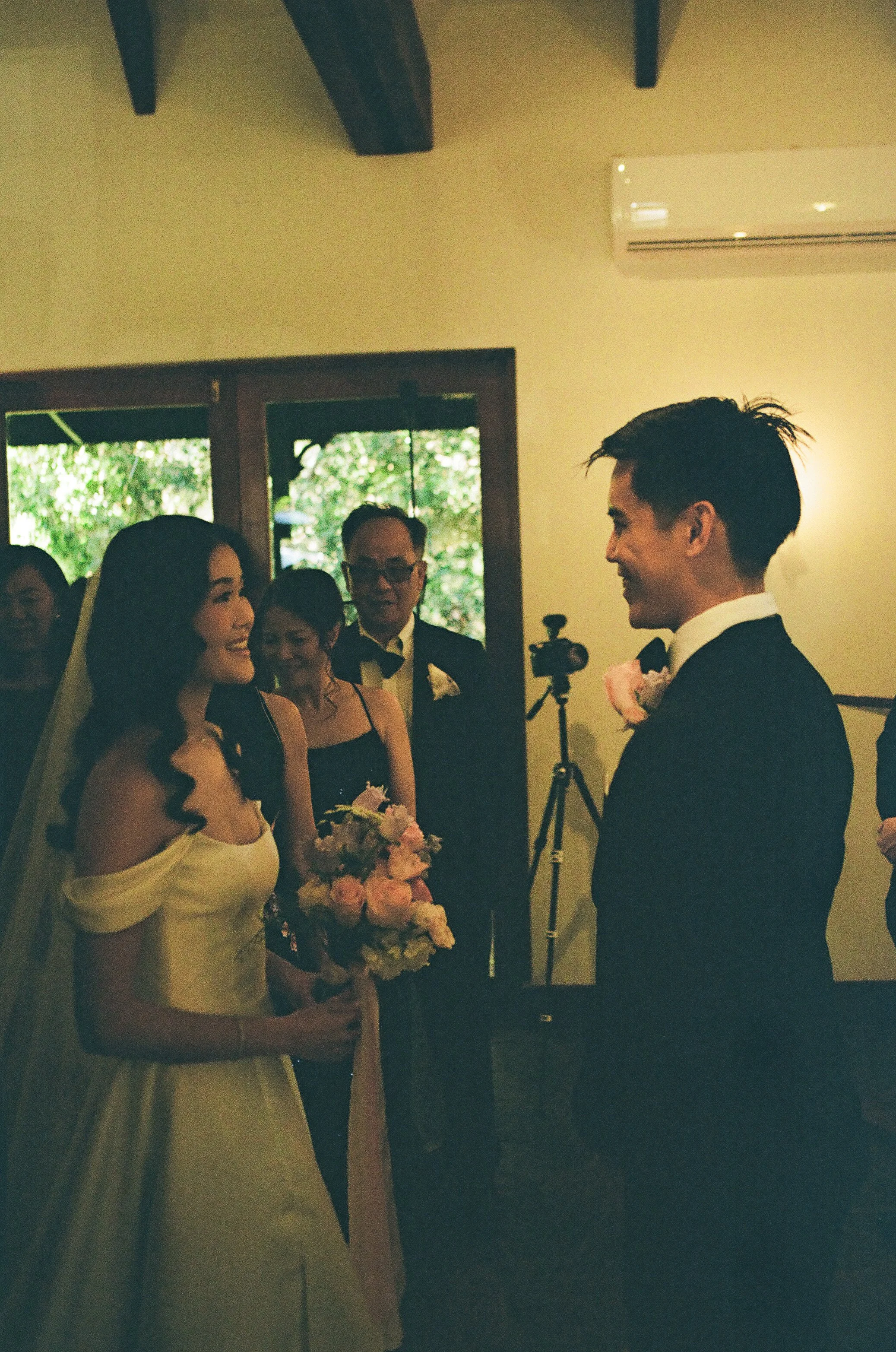 A bride in a yellow dress holding a bouquet of pink and white roses, smiling and looking at a groom in a black suit with a pink flower on his lapel, during a wedding ceremony, with friends and family gathered in a room with green outside views.