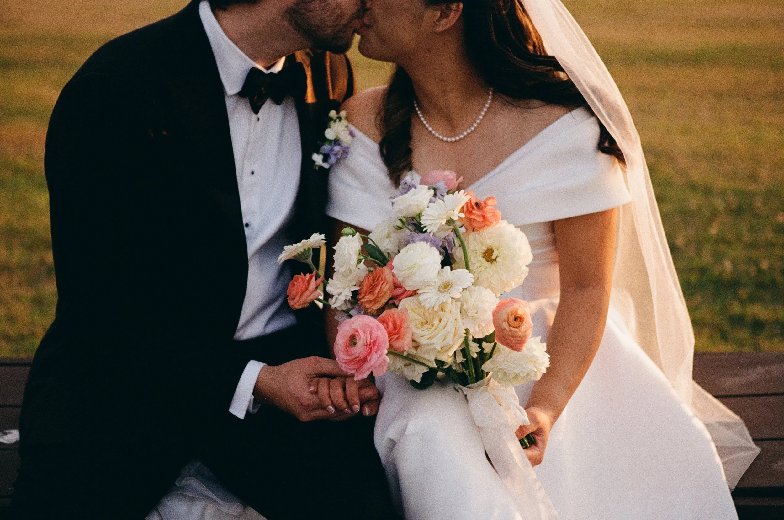 A newlywed couple sharing a kiss, with the bride holding a bouquet of flowers, sitting on a park bench during sunset.