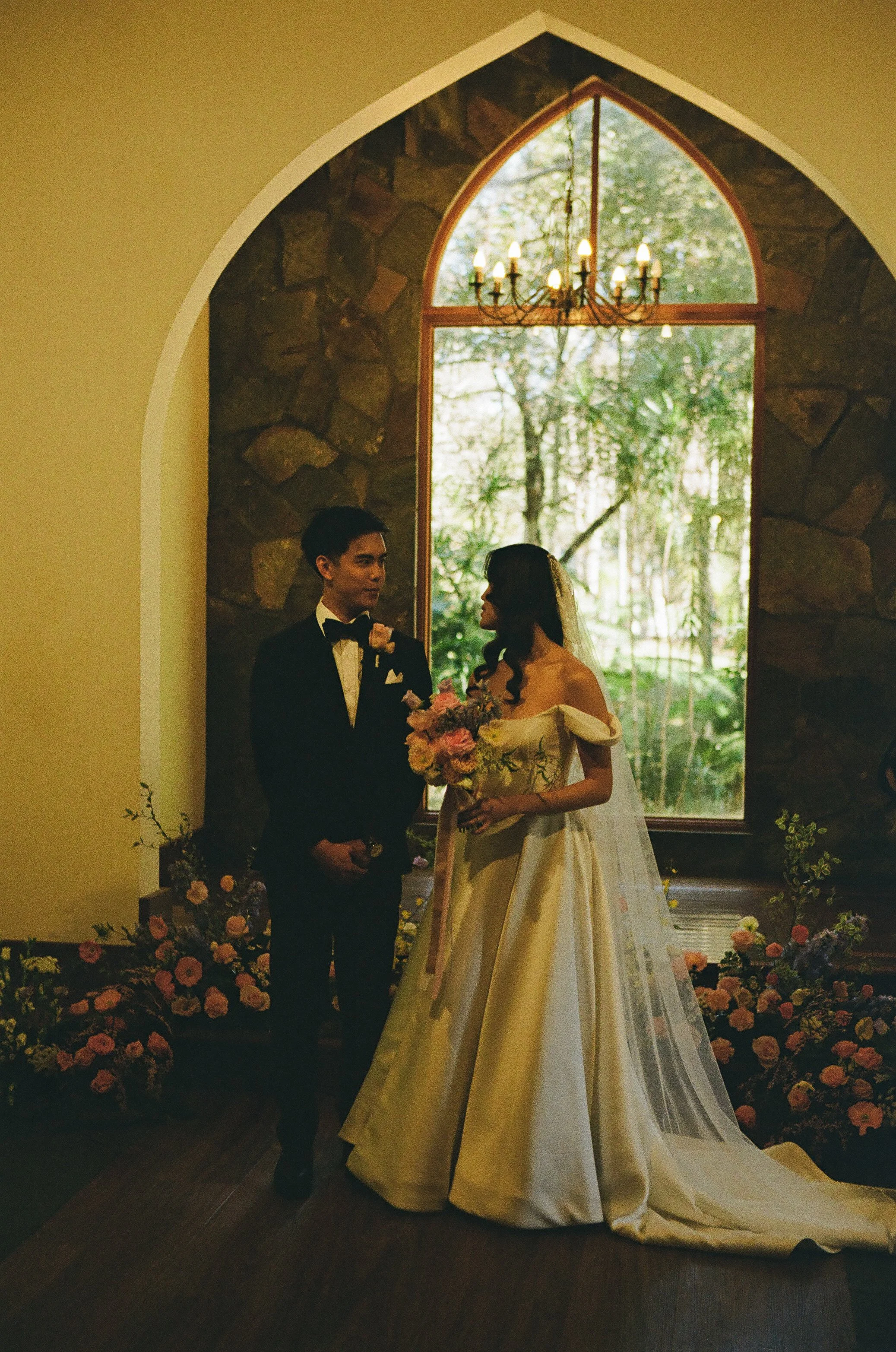 A bride and groom stand inside during their wedding ceremony, with the bride holding a bouquet of flowers, near a large arched window with a chandelier hanging above, and surrounded by floral arrangements.