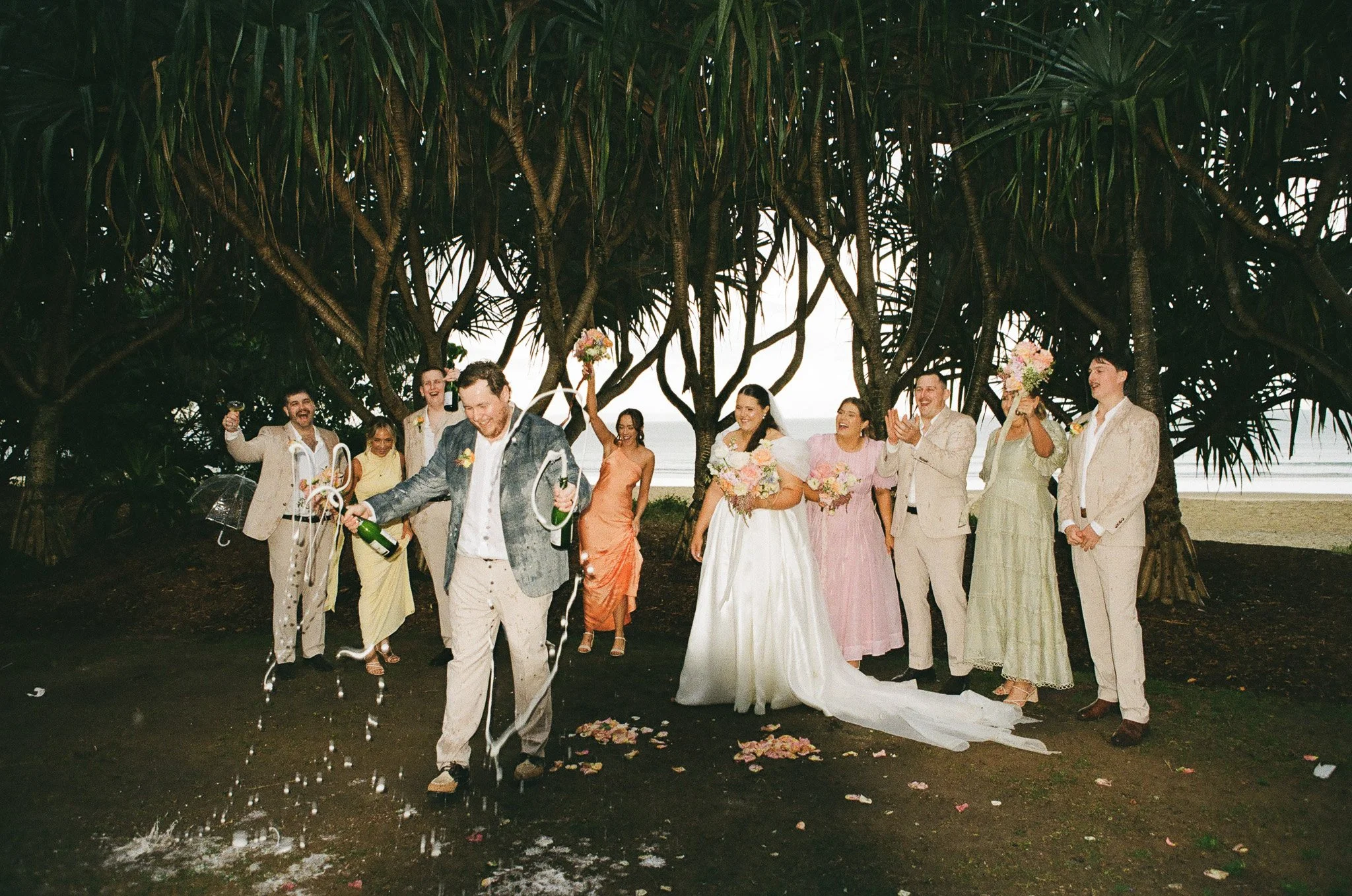 A wedding celebration outdoors with a bride in a white gown and veil, surrounded by smiling guests. Guests are wearing light-colored suits and dresses, and some are holding bouquets. One man in a suit is opening a champagne bottle while others cheer.