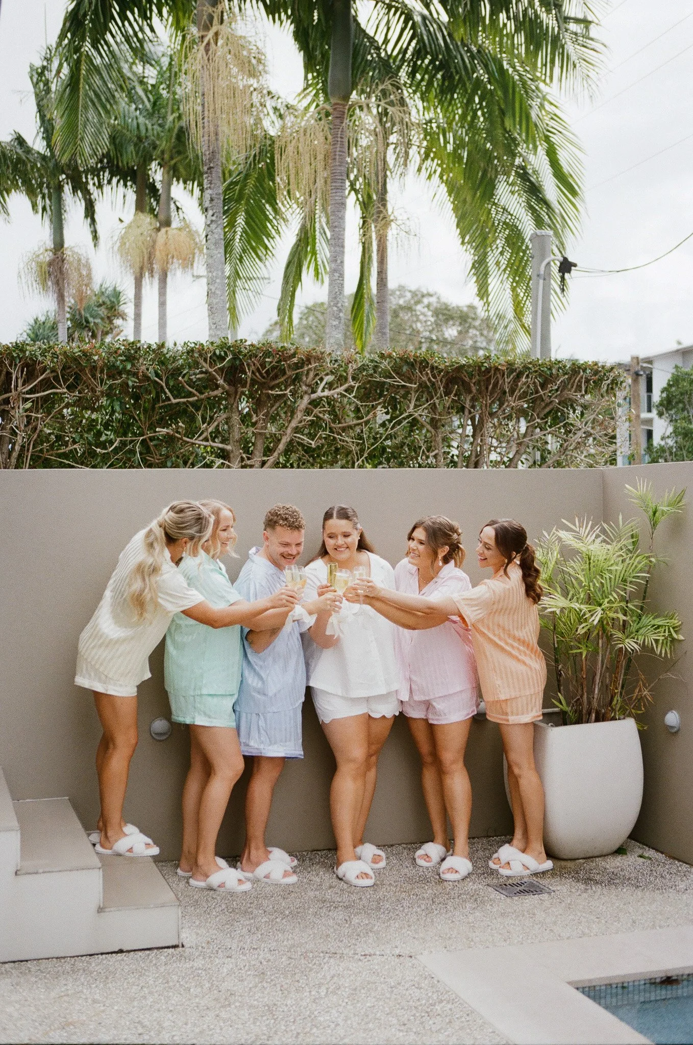 Six young women in pajamas, standing in a small outdoor patio, raising glasses of champagne in a toast. They are smiling and celebrating together, with a potted plant and a gray wall behind them.