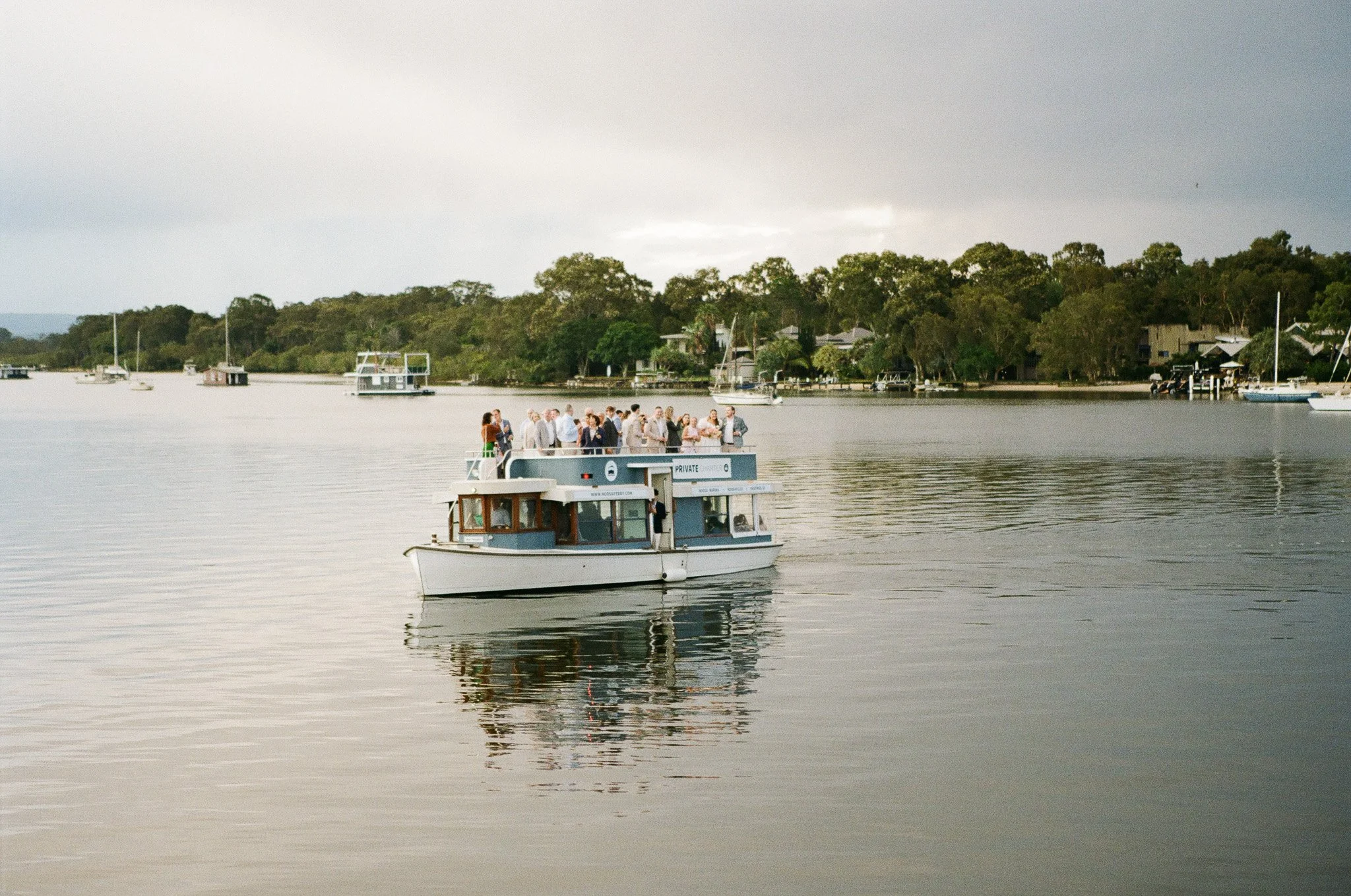 A group of people on a boat on calm water near a shoreline with trees and houses, under a cloudy sky.