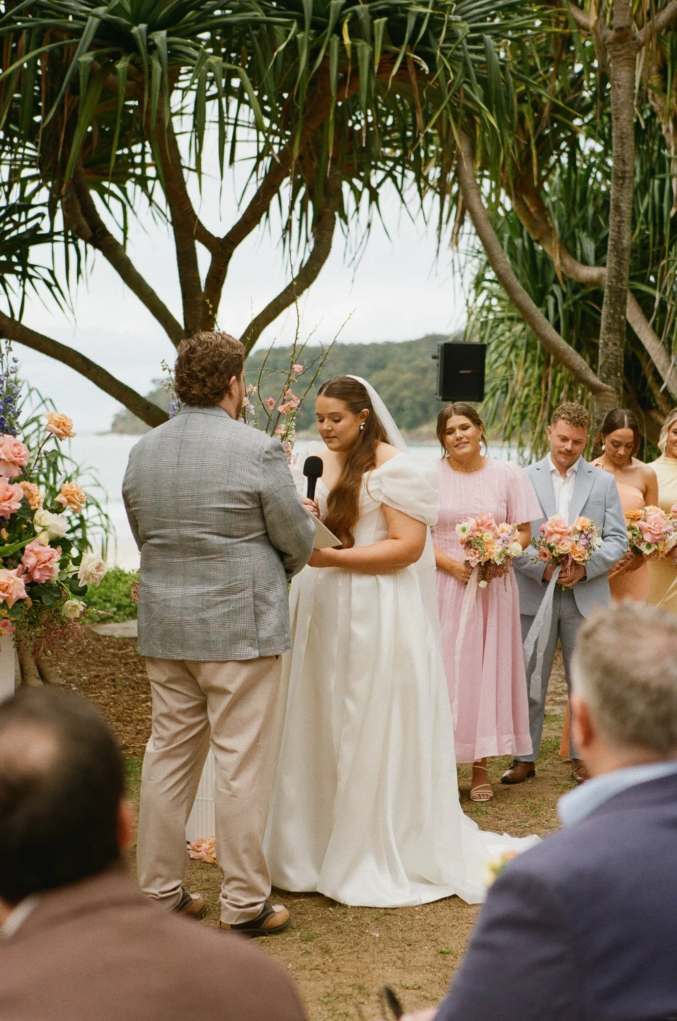 Wedding ceremony outdoors on a beach with a floral arch, with the bride and groom exchanging vows as a woman holds a microphone, surrounded by bridesmaids and groomsmen holding bouquets.