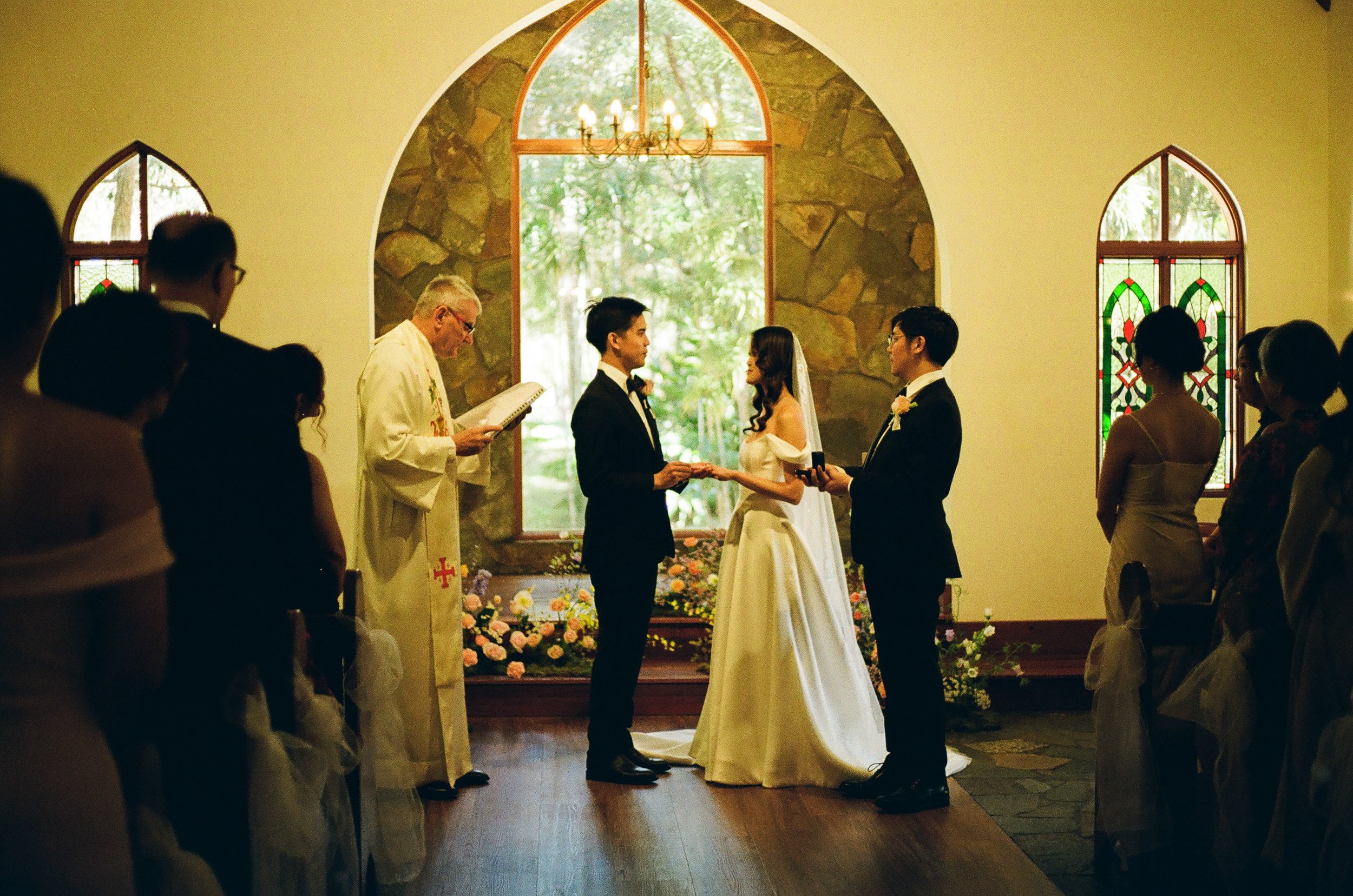 A wedding ceremony inside a church with a couple standing before an officiant and exchanging vows, surrounded by guests.