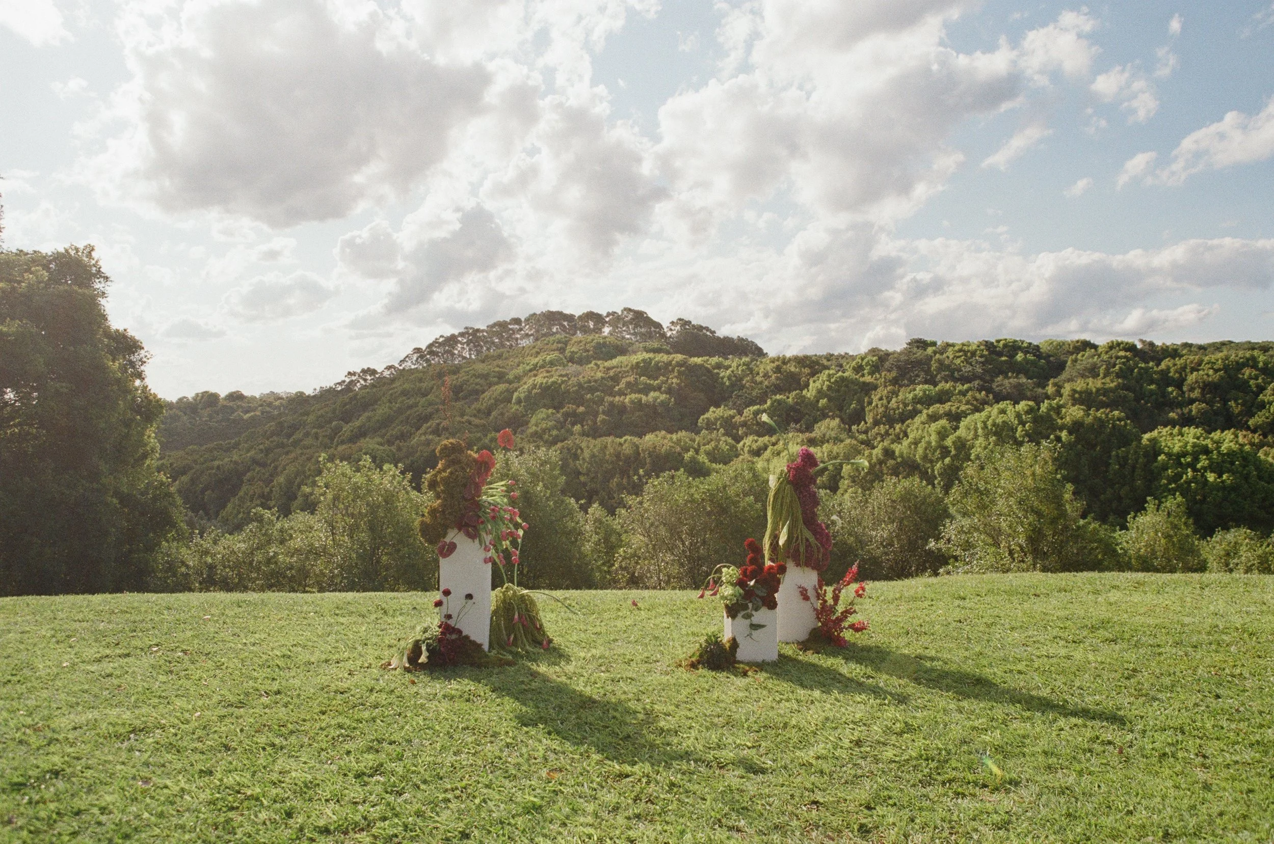 Outdoor scene with three white pedestals decorated with colorful flowers and greenery, set on a grassy field with a backdrop of green hills and a partly cloudy sky.